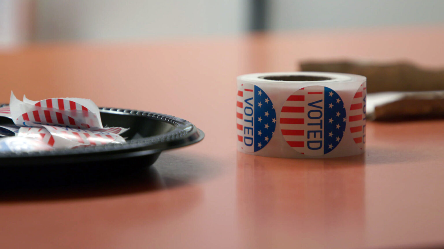 A roll of stickers with a stars-and-stripes graphic and the words I Voted sits on a table next to a disposable plastic plate filled with individual stickers.