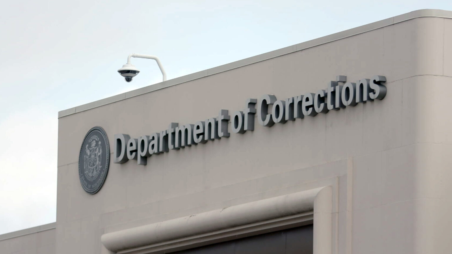 An entrance sign near the top of a  concrete-masonry building includes a Wisconsin state seal and letters spelling out Department of Corrections, with a surveillance camera mounted from the edge of the roof.