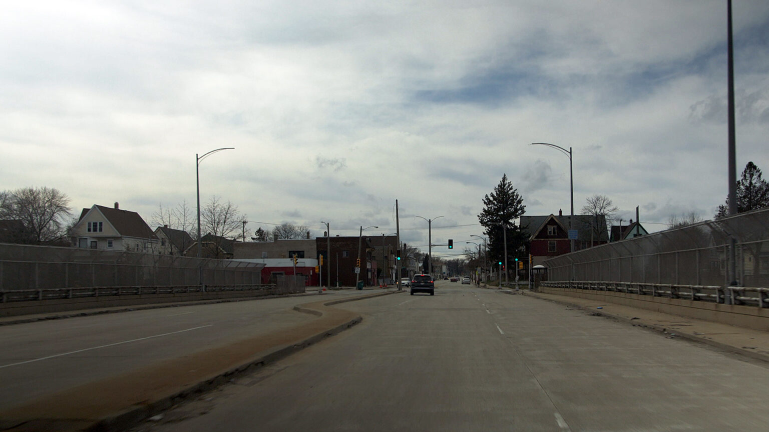 A perspective from the front window of a vehicle shows a divided street on a bridge, with other vehicles, street lights, trees and buildings in the background.