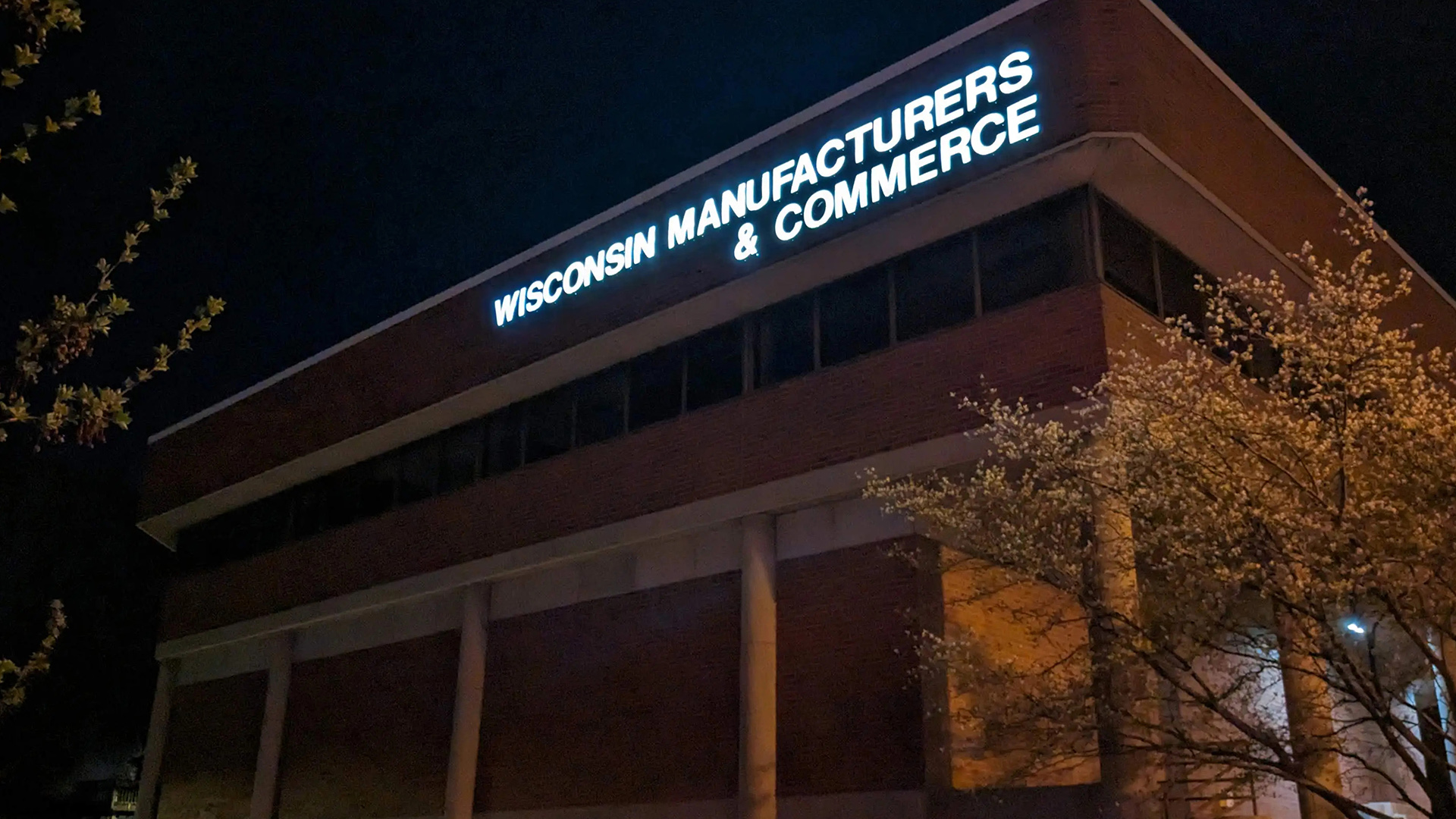 Lighted letters reading "Wisconsin Manufacturers & Commerce" are placed at the top of a building with concrete pillars, brick walls and one row of windows, with trees in the foreground.