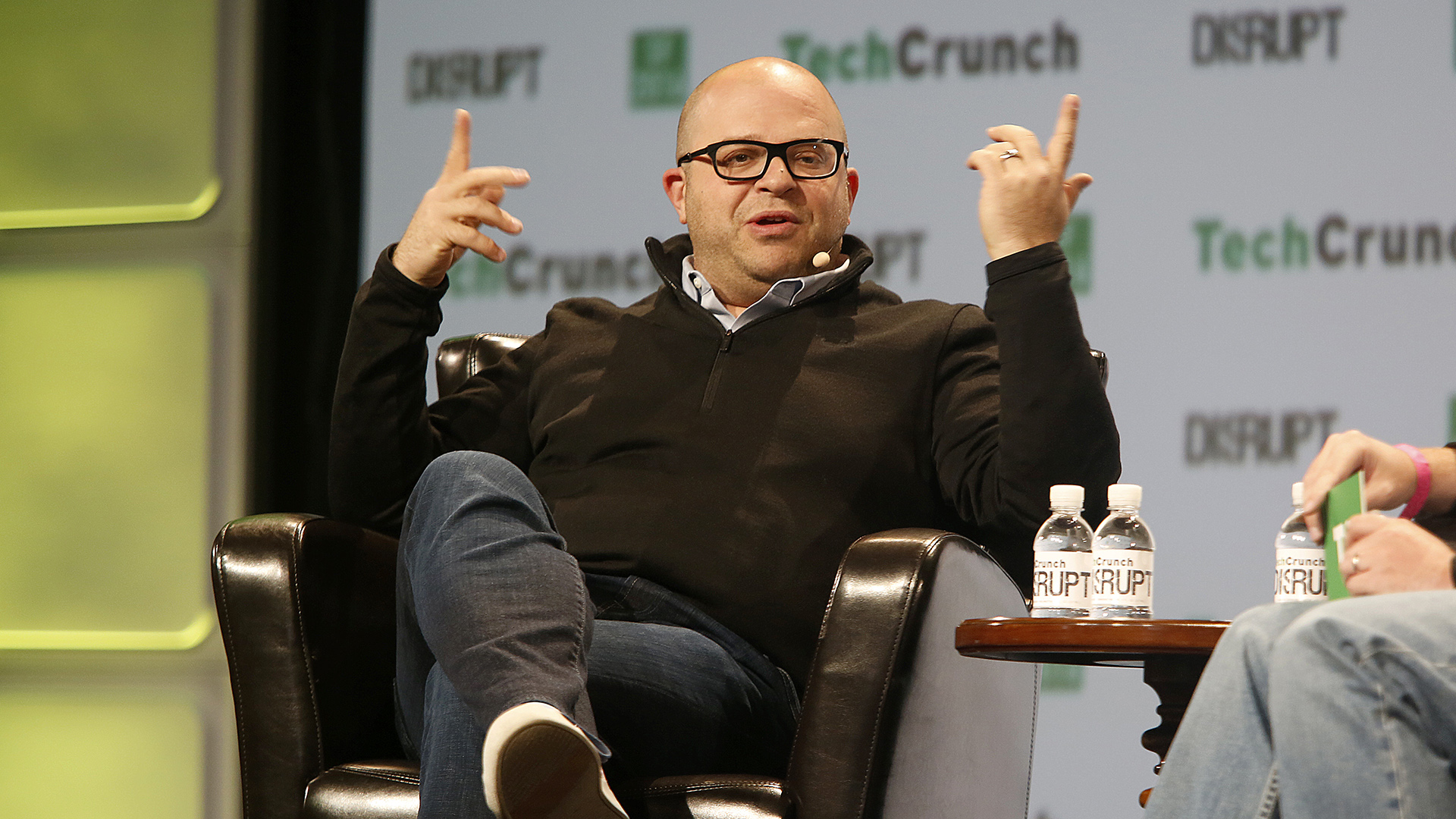 Jeff Lawson sits in an arm chair and gestures with both hands while speaking to another person seated by his side, in a room with a logo backdrop with wordmarks that read "TechCrunch" and "DISRUPT."