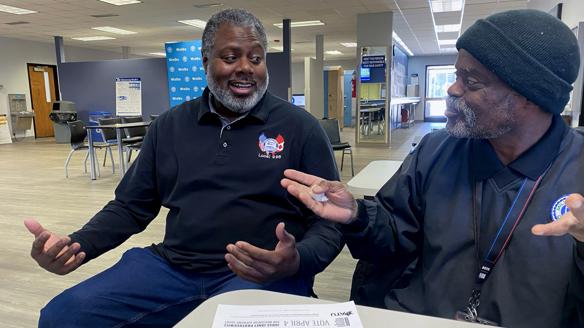 Donnell Shorter sits at the corner of a folding table and gestures with both hands while speaking to a man seated to his side and is also gesturing with both hands, in a room with more tables, cubicle dividers and lockers in the background.