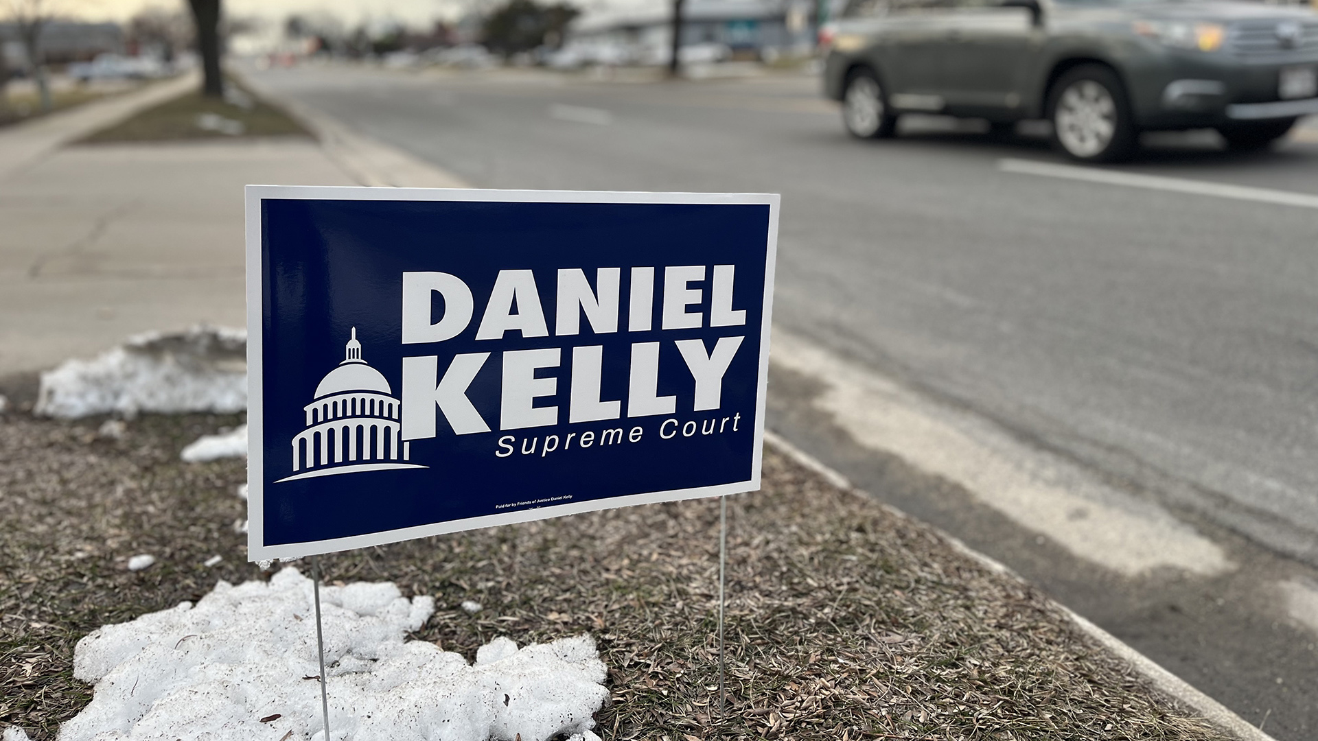 A campaign yard sign with a Capitol dome graphic and the words "Daniel Kelly" and "Supreme Court" stands in a grass median between a street and sidewalk, with a driveway, driving car, buildings and trees in the background.