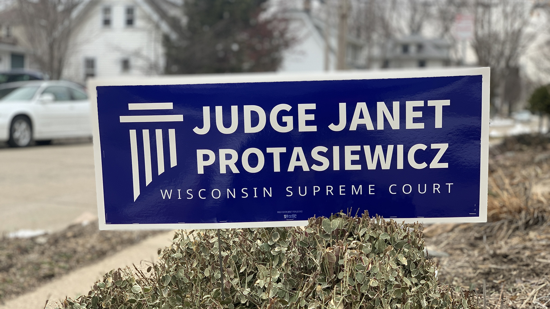 A campaign yard sign with a pillar graphic and the words "Judge Janet Protasiewicz" and "Wisconsin Supreme Court" stands behind a shrub in a yard next to a sidewalk, with a street, parked car, houses and trees in the background.