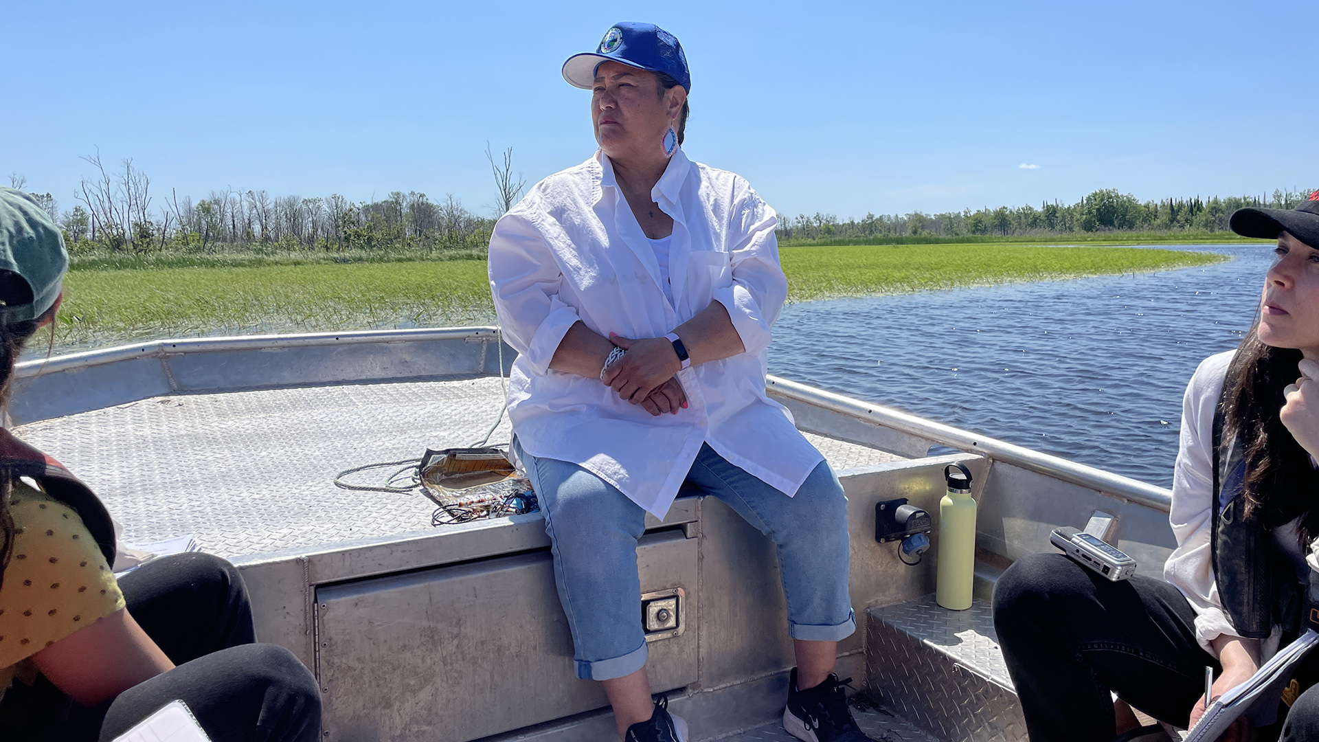 Edith Leoso sits on the bow of a metal boat and looks over the water, with two other people seated to the side and with wild rice beds and trees in the background.