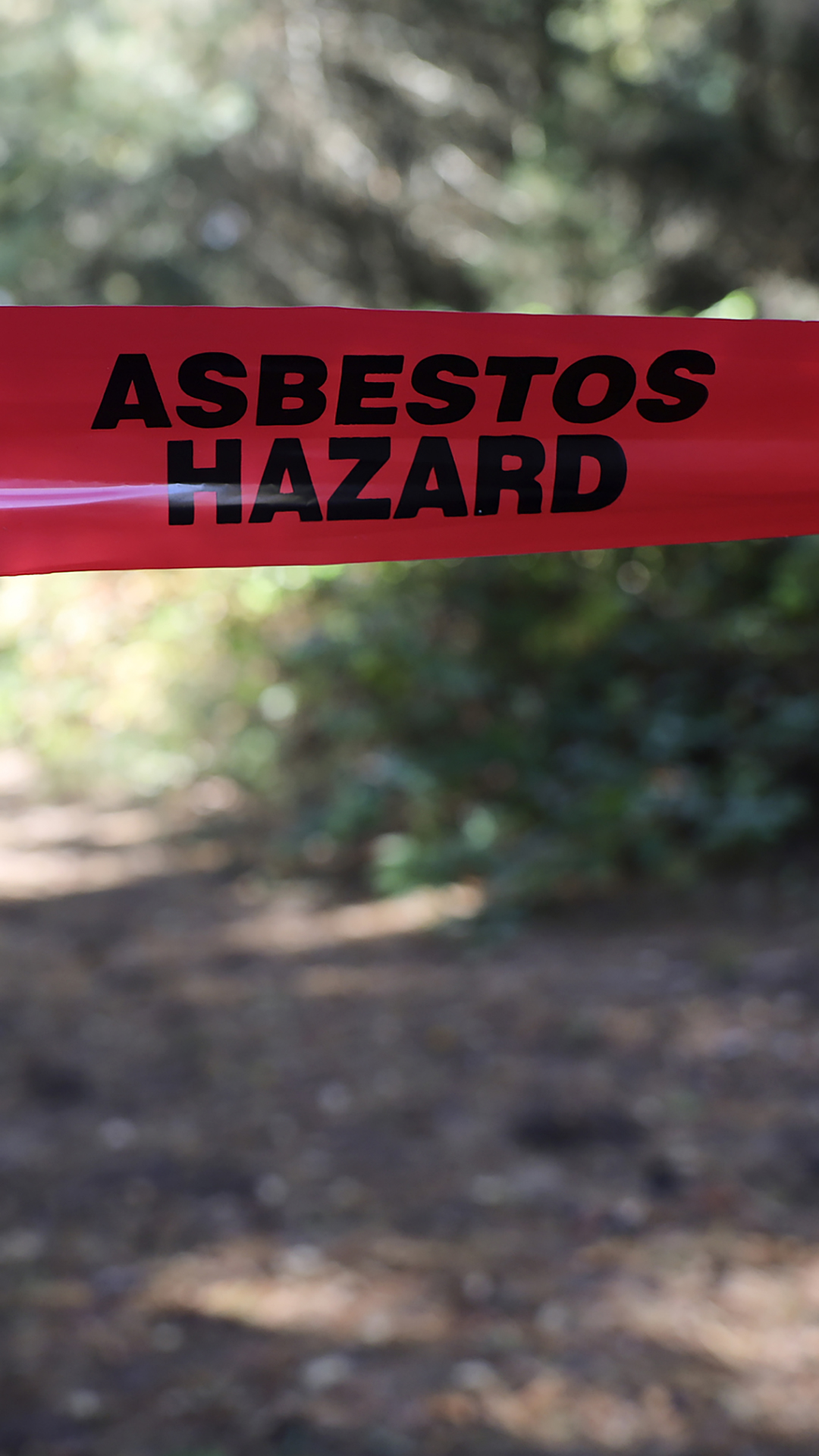 Plastic tape bearing the words "Asbestos Hazard" crosses a path in a wooded area, with sunlight trees in the background.
