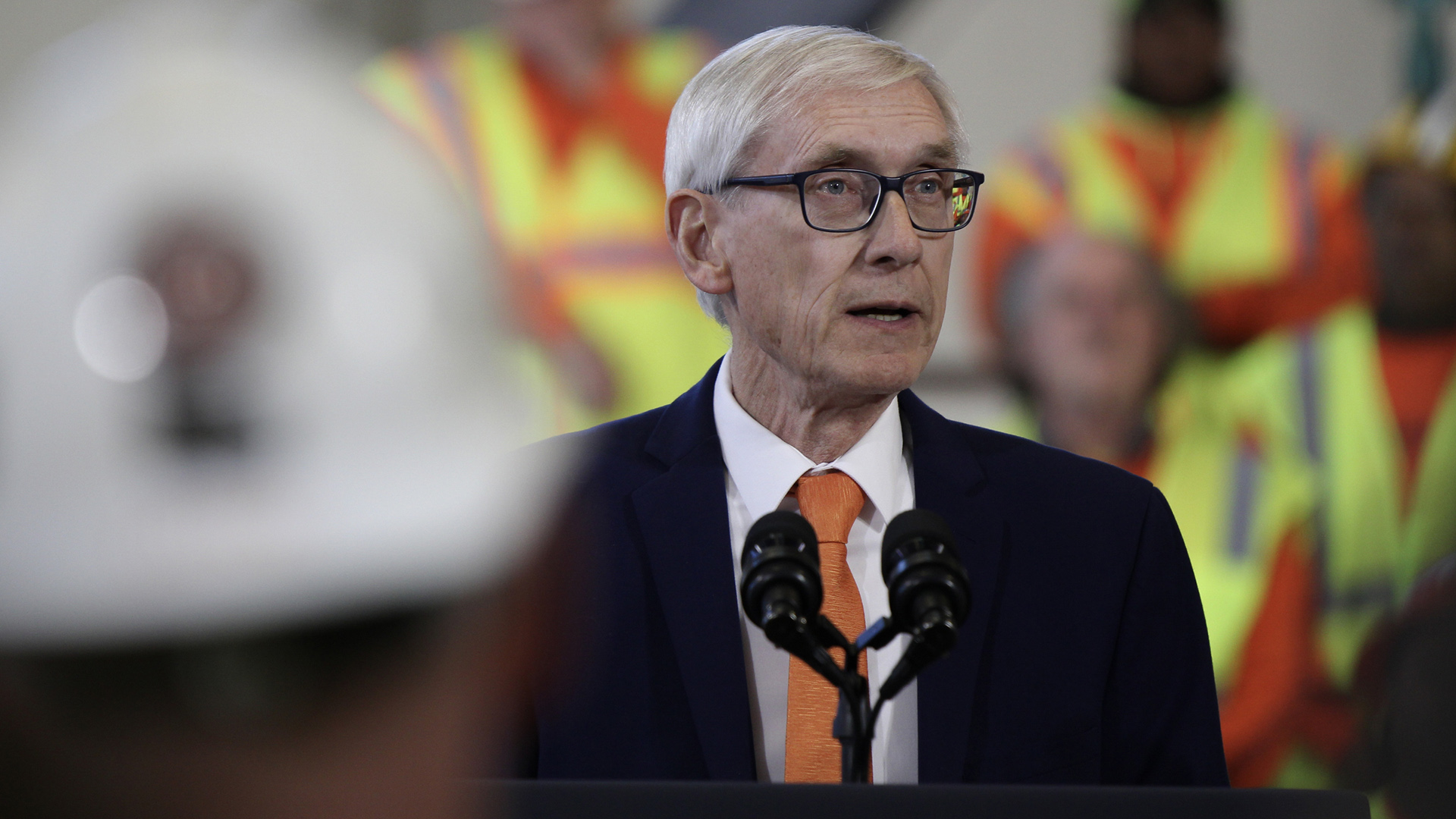 Tony Evers speaks into a pair of microphones mounted on a podium with out-of-focus people wearing reflective safety vests standing in the background and an out-of-focus person wearing a construction helmet in the foreground.