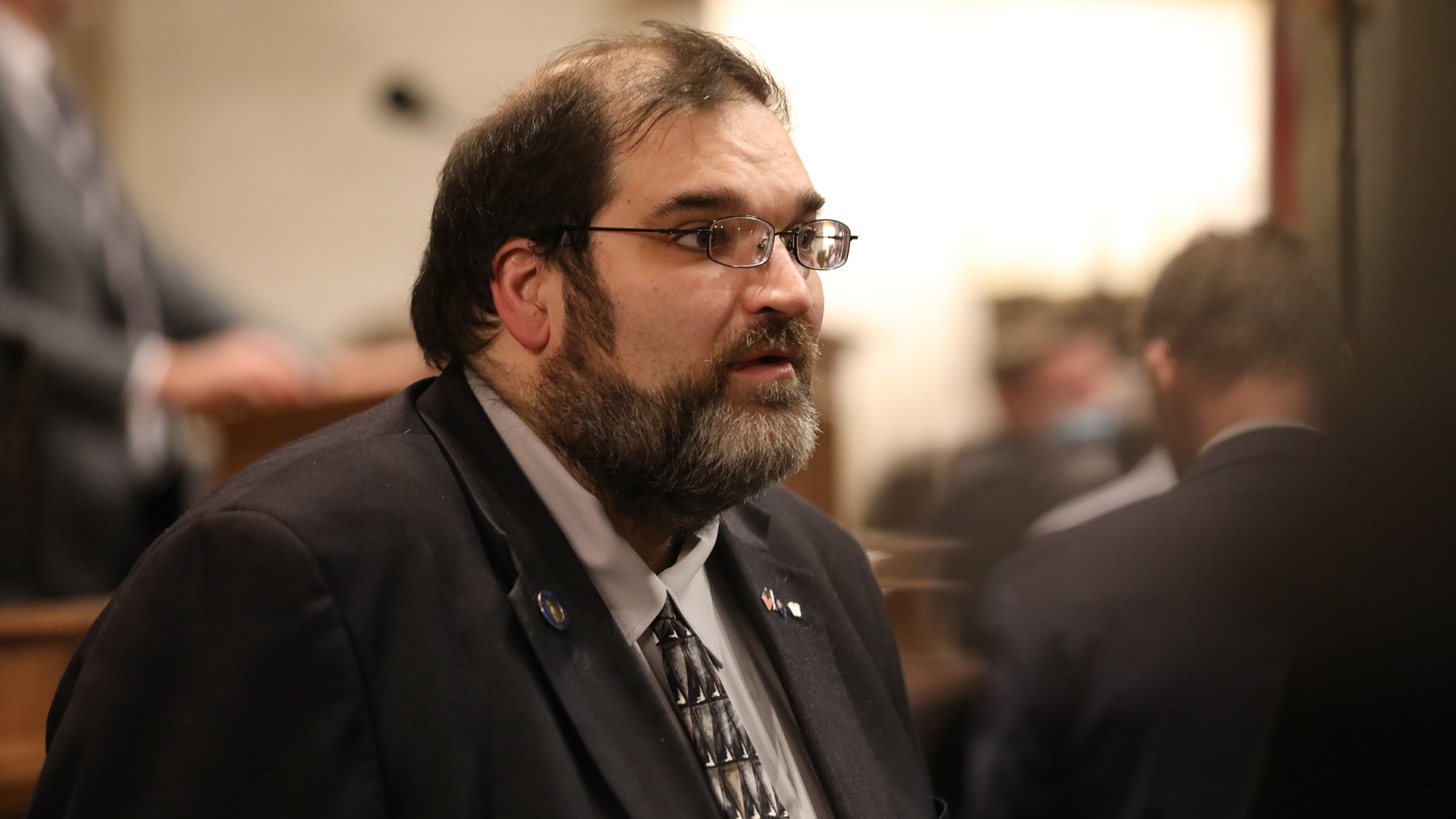 Andre Jacque stands in a room and speaks, with other out-of-focus people standing in the background.