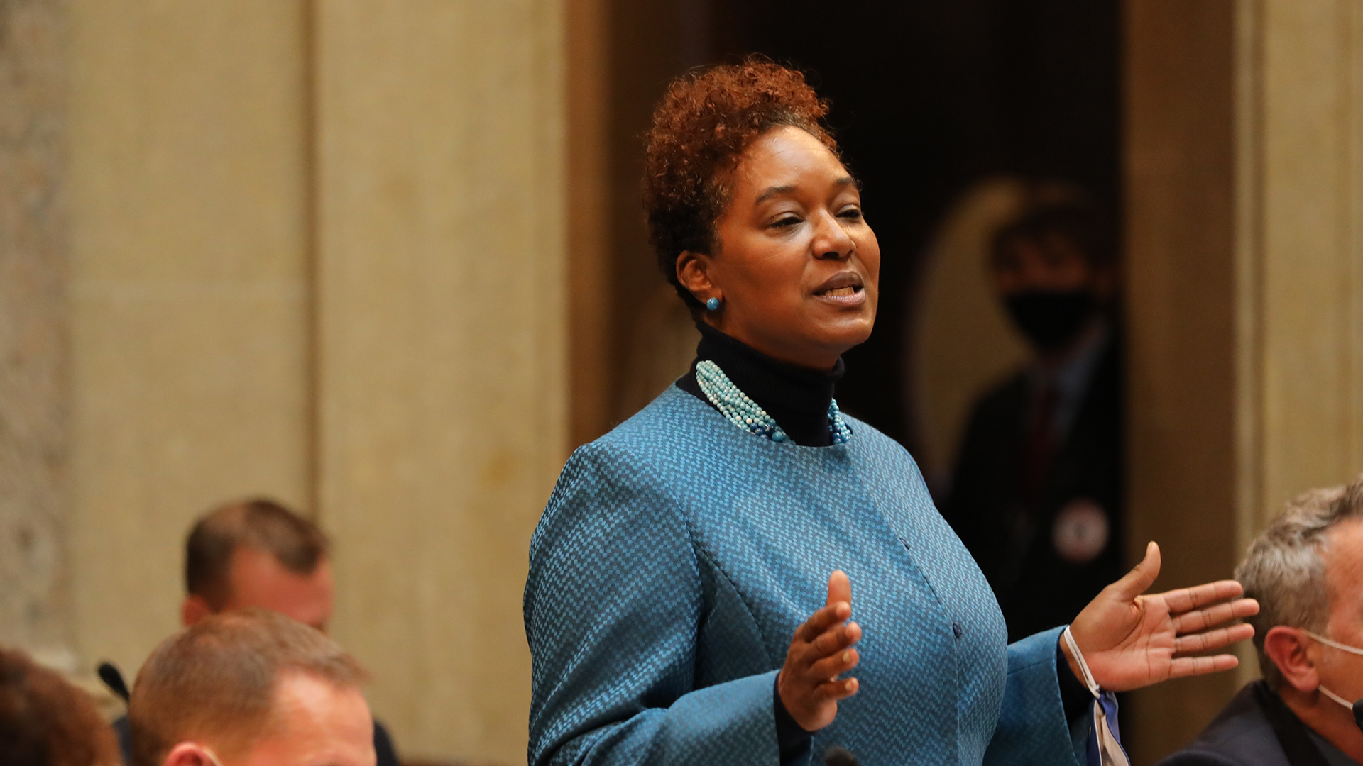 Lena Taylor stands and speaks while gesturing with both hands while other people are seated around her in a legislative chamber.