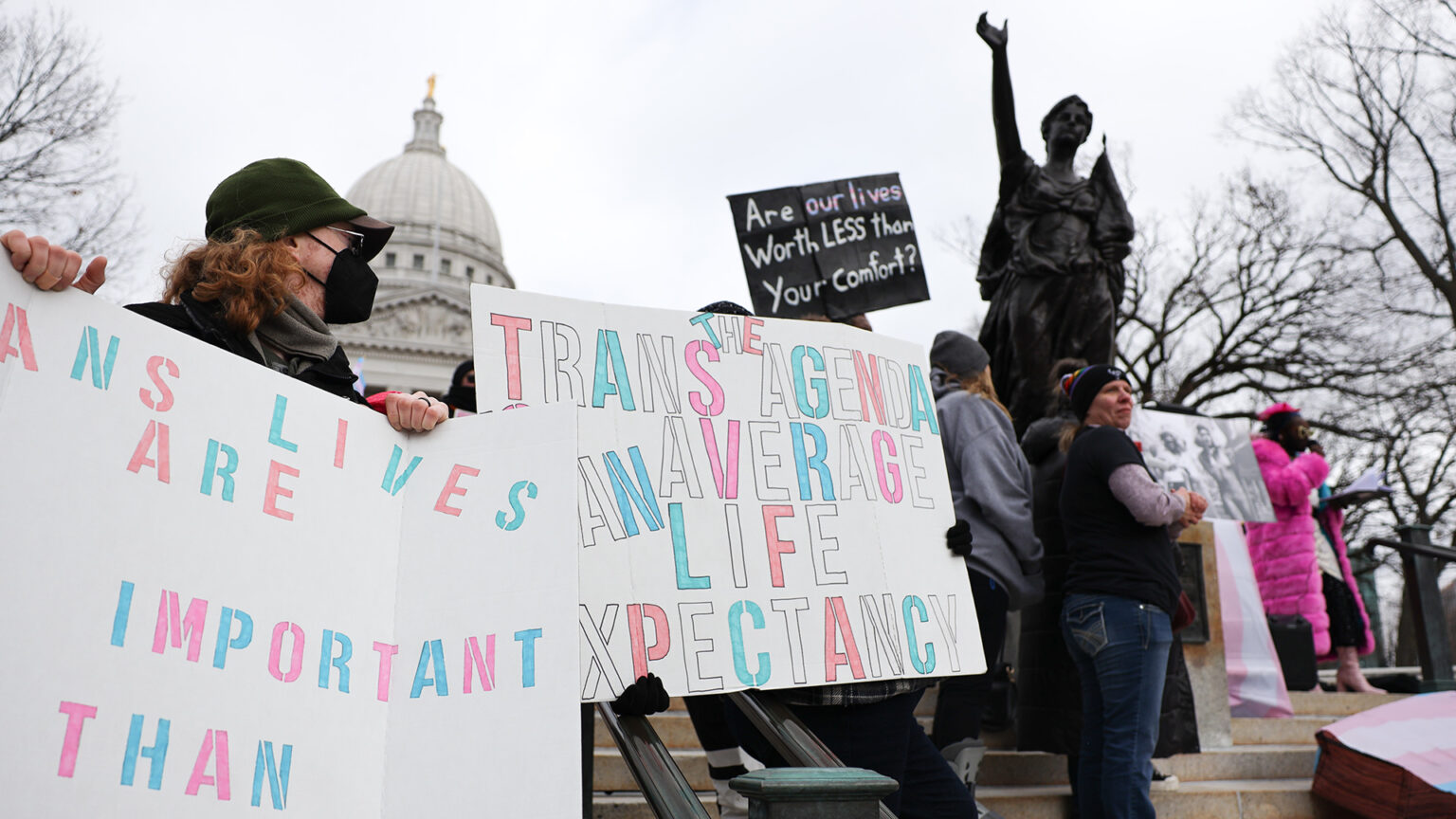 A group of people stand on steps around a statue and hold signs, with trees and the dome of the Wisconsin State Capitol in the background.