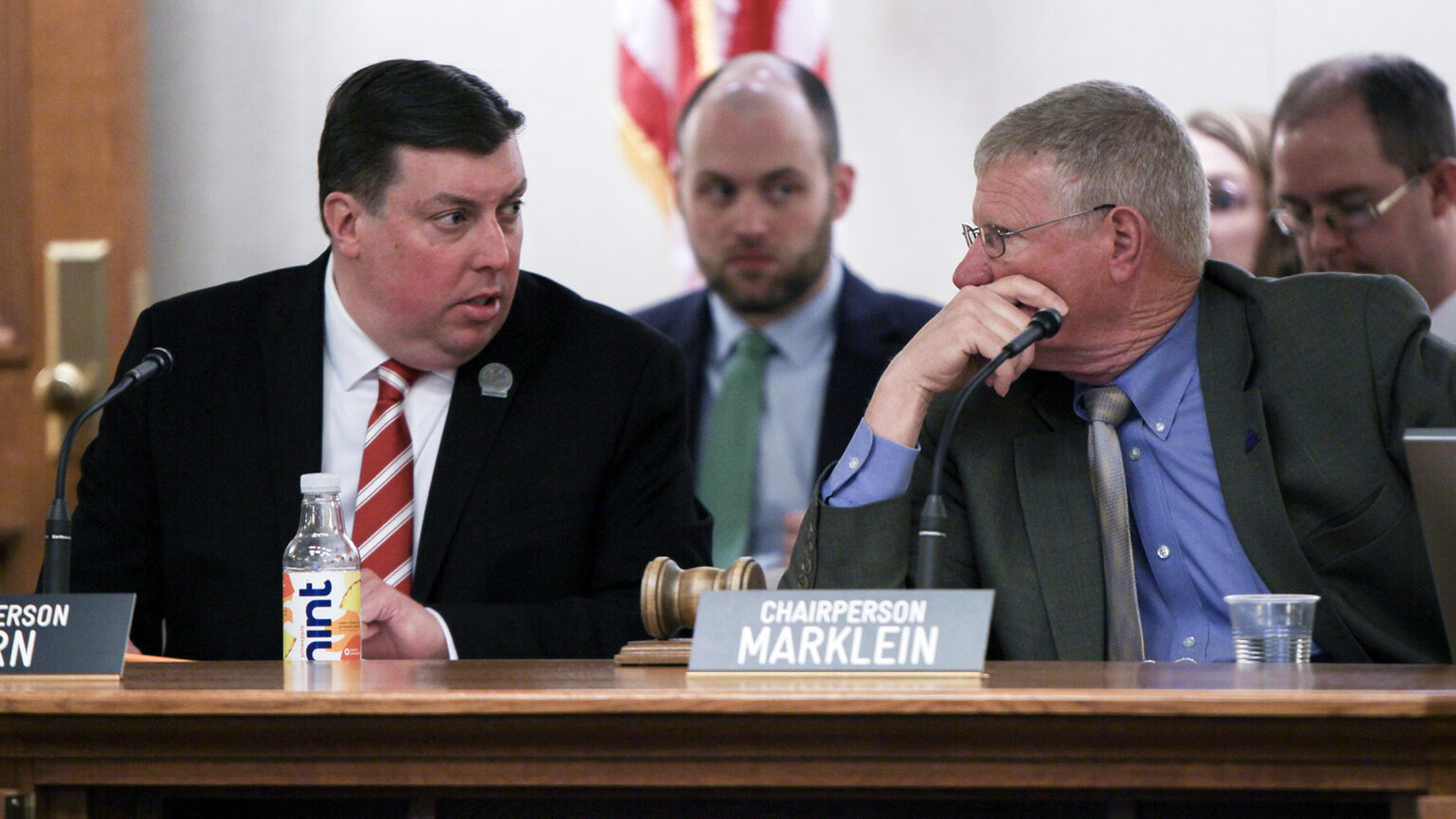 Mark Born and Howard Marklein sit at a legislative dais with microphones and signs with their names, speaking to each other and with other people seated in the background.