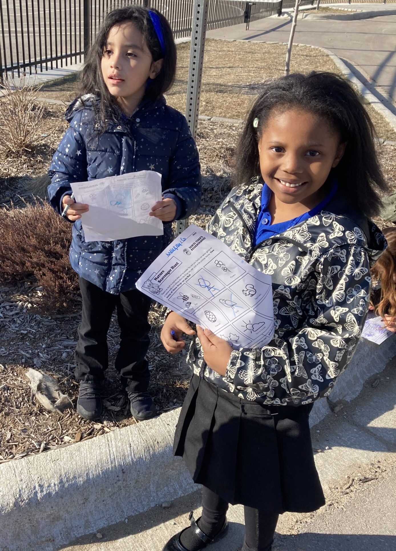 Two children stand outside, each with a piece of paper in hand. They are on a scavenger hunt.