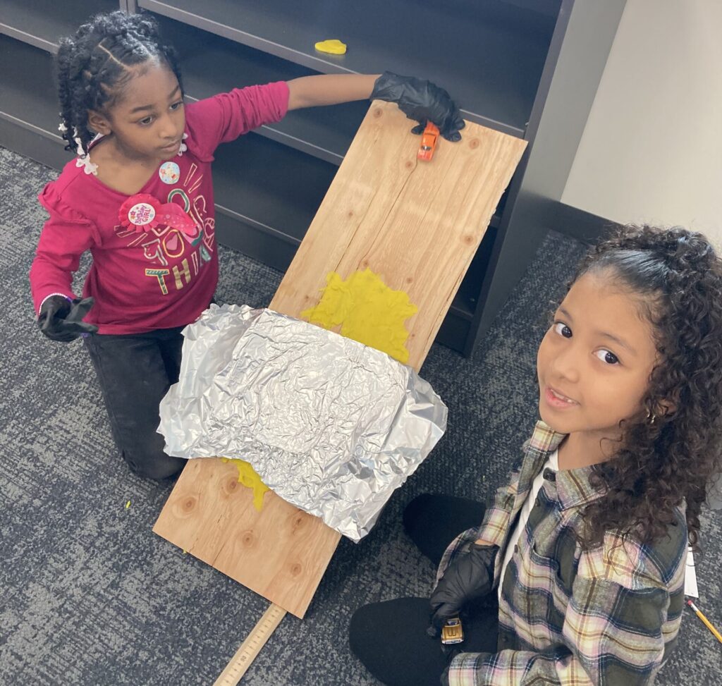 Two elementary aged children sit on the floor next to a science project. One child holds a car at the top of a slanted board, ready to release the car. The other child turns and smiles at the camera.