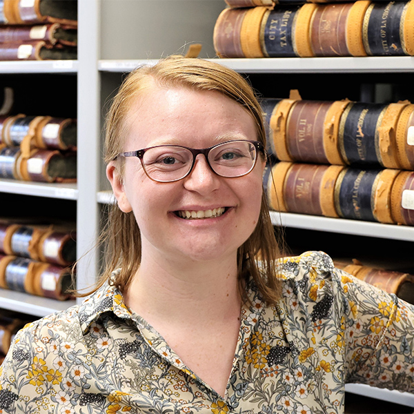 Woman wearing glasses and smiling in front of shelving holding tax records.