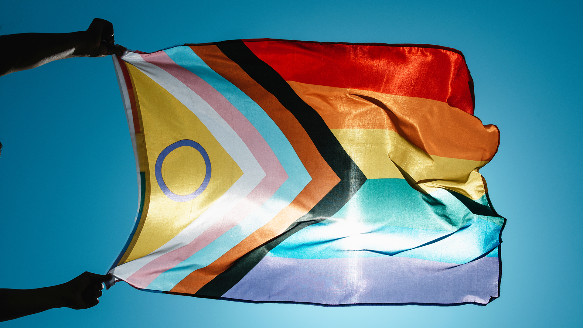 An LGBTQIA+ Progressive Pride rainbow flag is held up by two hands against a blue sky with light shining through the flag.