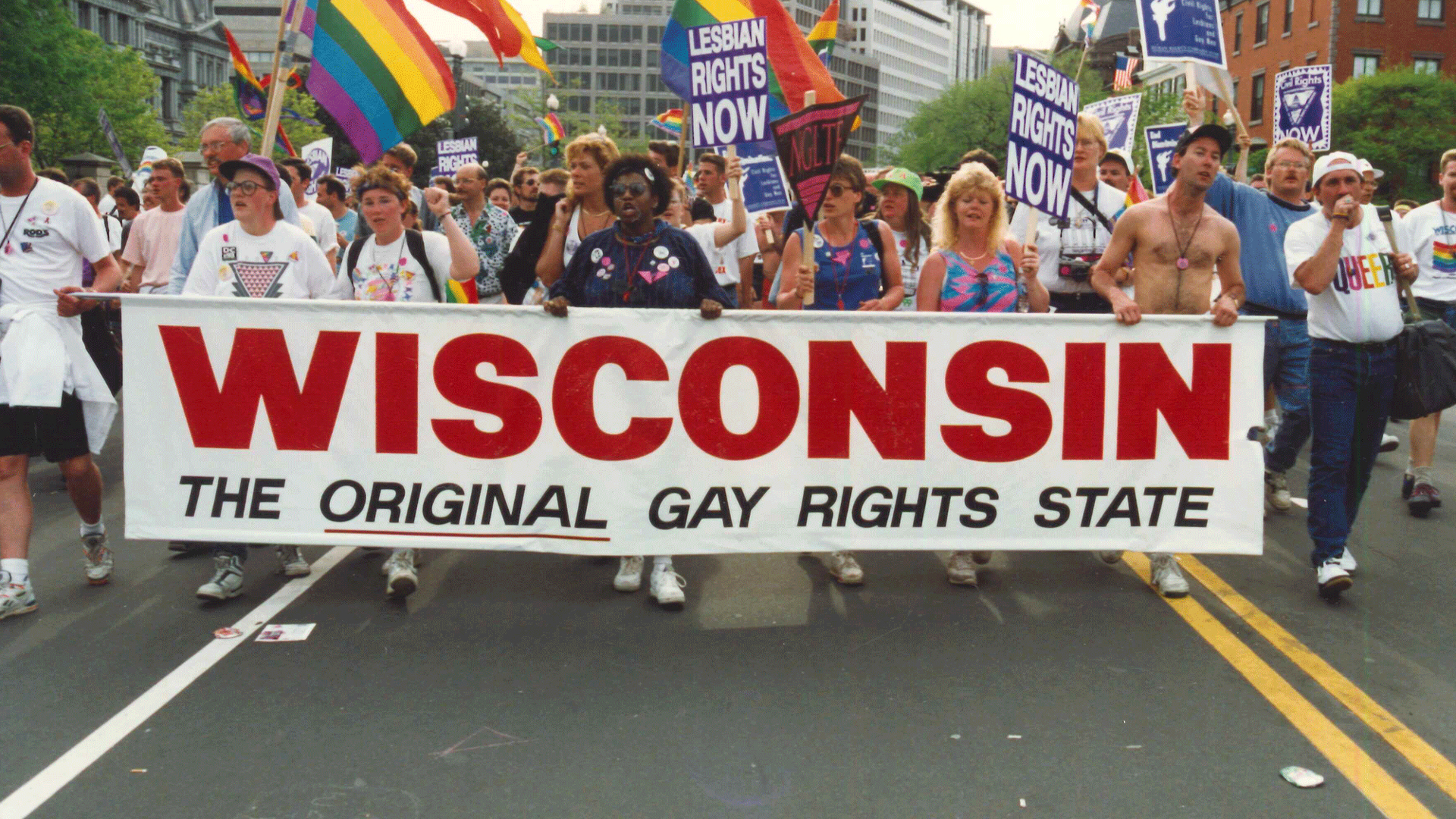 A gay rights pride march in 1982, a large group of people lead with a banner reading "WISCONSIN, the original gay rights state" followed by rainbow Pride flags and LGBTQ+ rights signs.