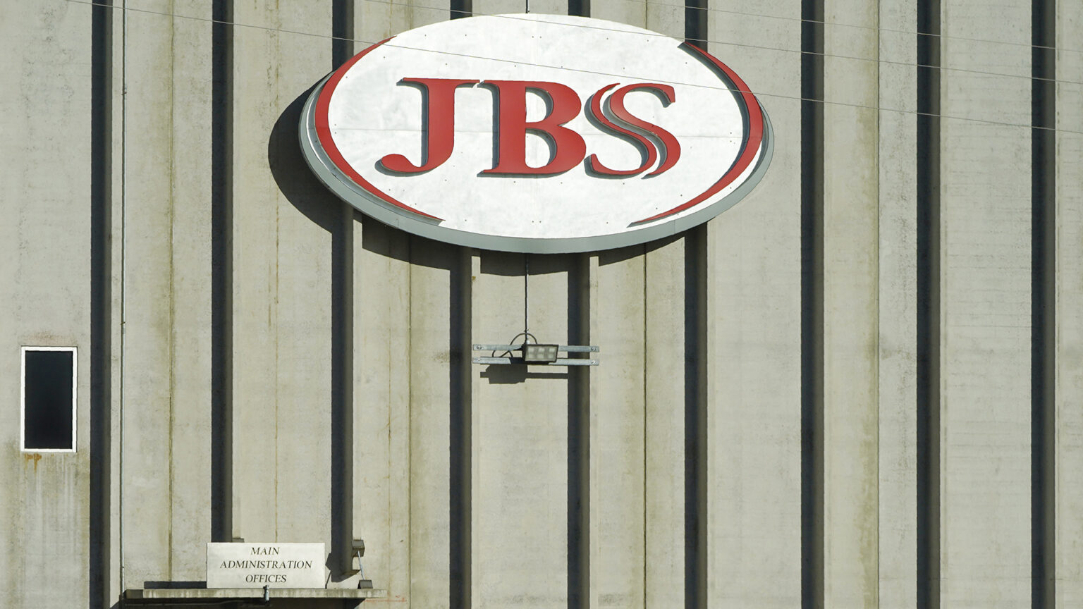 A sign with a wordmark logo that reads JBS is fixed to the wall of a multi-story building with concrete and metal walls, with a sign atop a door reading Main Administration Offices.