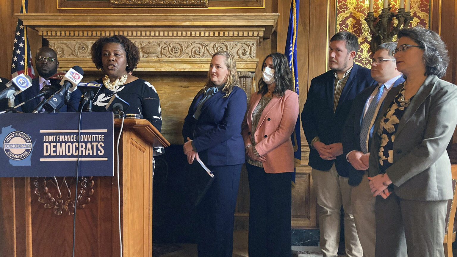 LaTonya Johnson stands at a podium with a sign with the words Joint Finance Committee Democrats and speaks into multiple microphones, with other people standing behind her in a room with gilt toile wallpaper, a carved wood mantel for a fireplace, and the U.S. and Wisconsin flags.