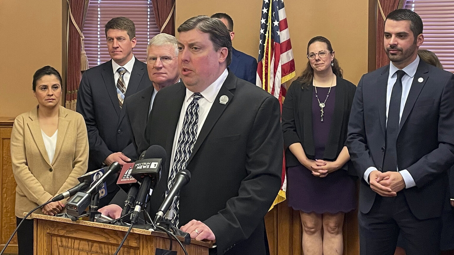 Mark Born stands behind a wood podium affixed with multiple microphones and speaks, while multiple other people stand behind him in a room with a U.S. flag and windows with lowered and closed blinds.