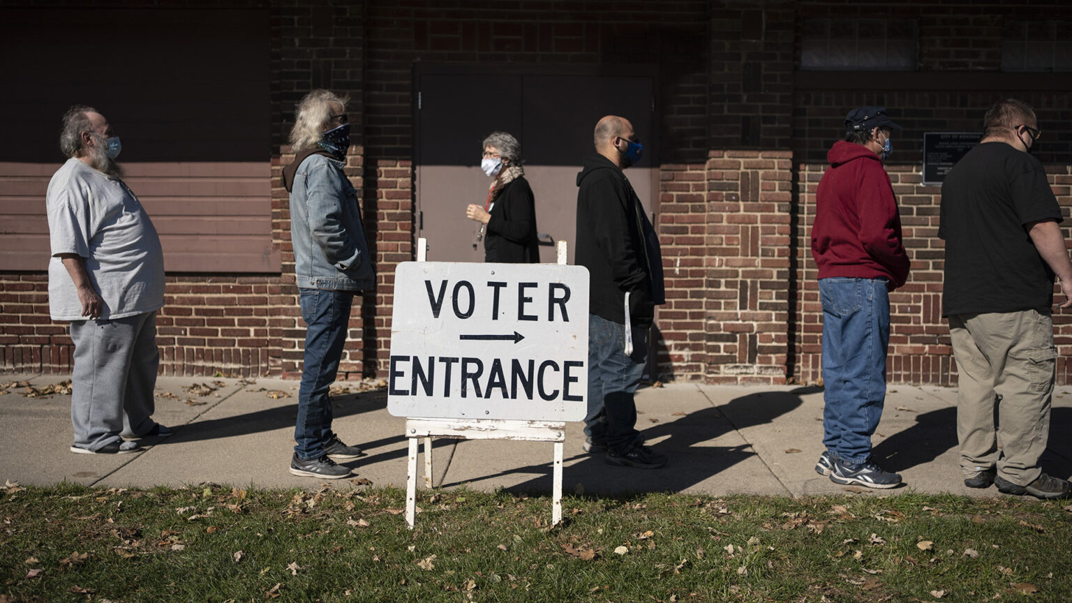 People stand in line on a sidewalk next to a building with brick walls, with a sandwich board sign in the foreground reading Voter Entrance with an arrow pointing to the right.