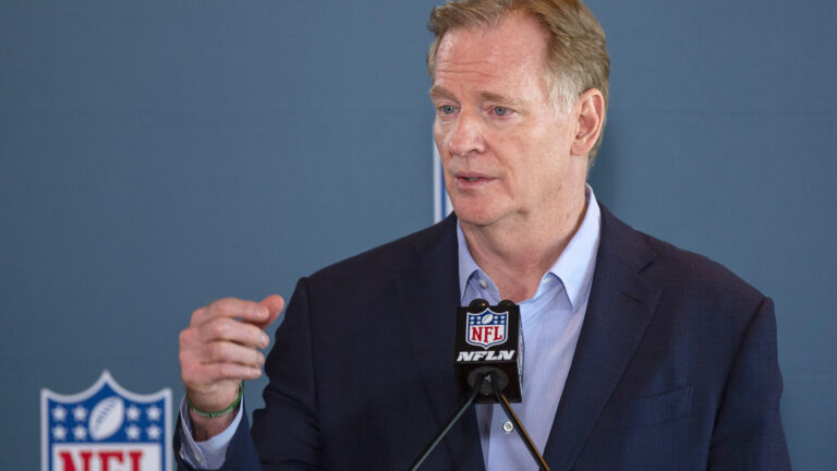 Roger Goodell stands and gestures with his right hand while speaking into a microphone with the NFL logo and the letters NFLN in front of a wall featuring multiple NFL logos.