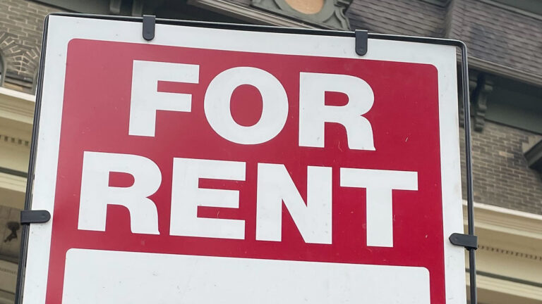 A temporary yard sign with the words For Rent on it stands in front of a dwelling with brick walls and a mansard roof.