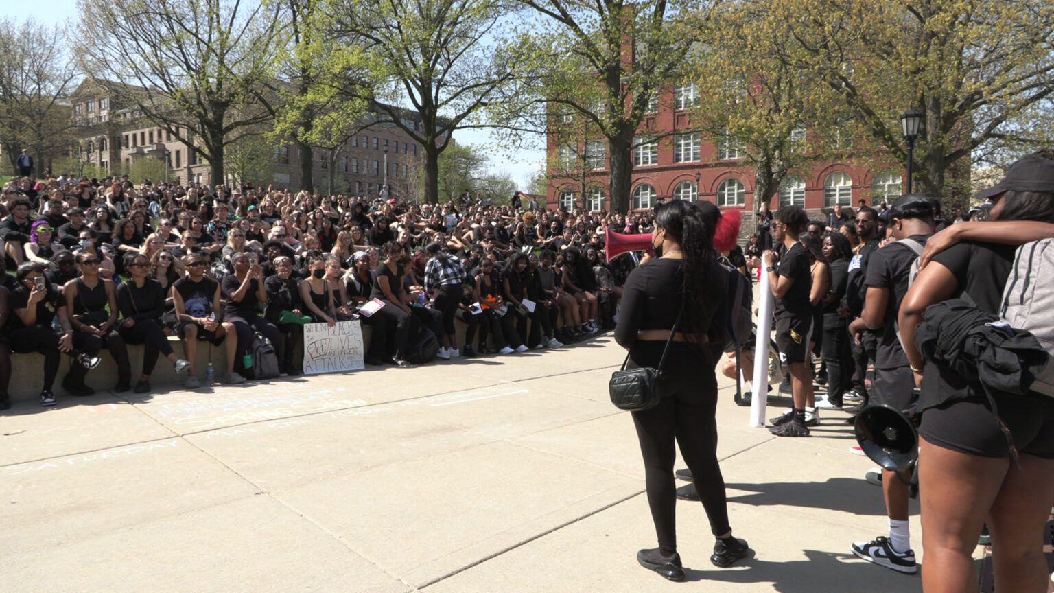 A large group of people sit at the base of a hill and listen to a speaker holding a megaphone, with more people standing to their side, with multi-story brick buildings and blooming trees in the background.