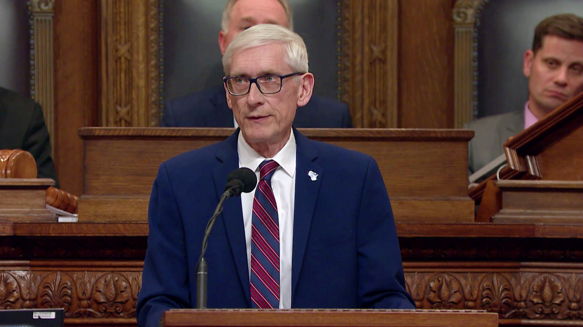 Tony Evers stands at a podium and speaks into a microphone, with other people seated behind him in high-backed leather chairs at a wood dais with ornamental carvings.