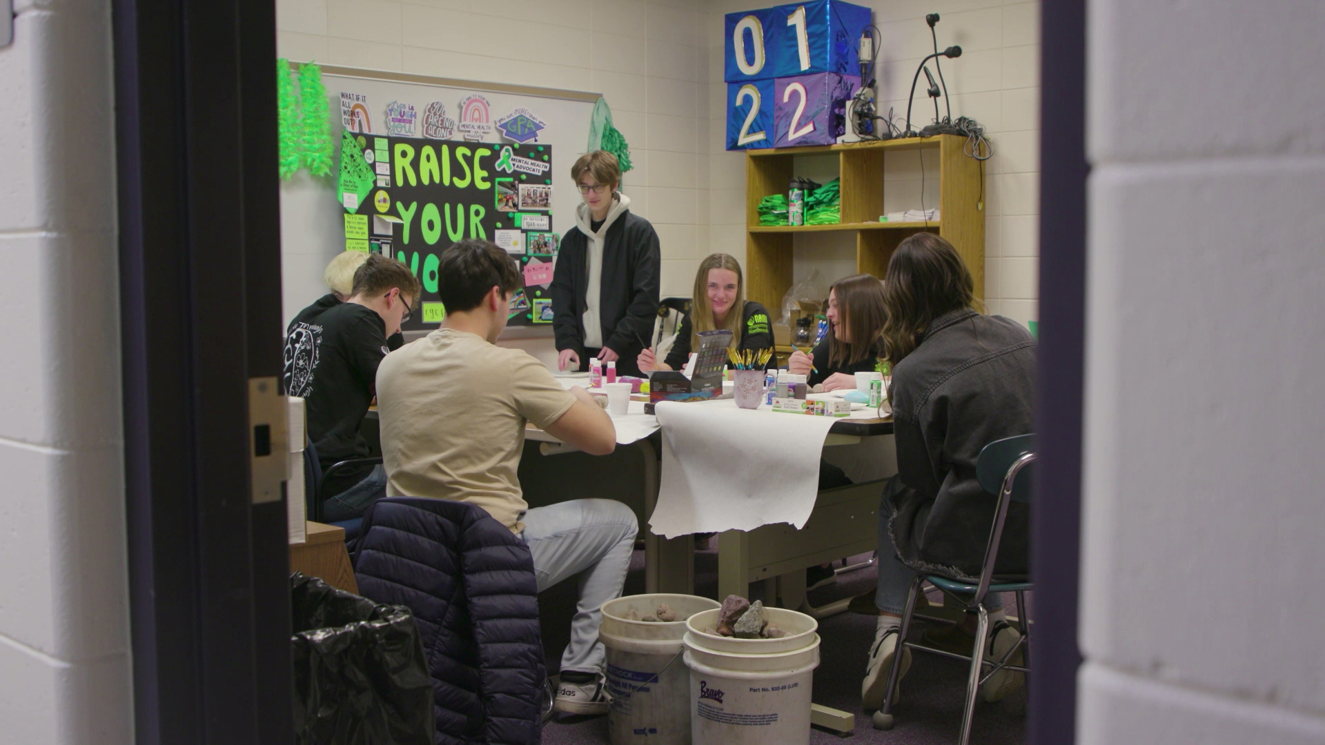 Multiple students sit around a table and work on a painting project in a room with painted concrete-block walls, shelves, a whiteboard and collage-style poster with the words "Raise Your Voice."