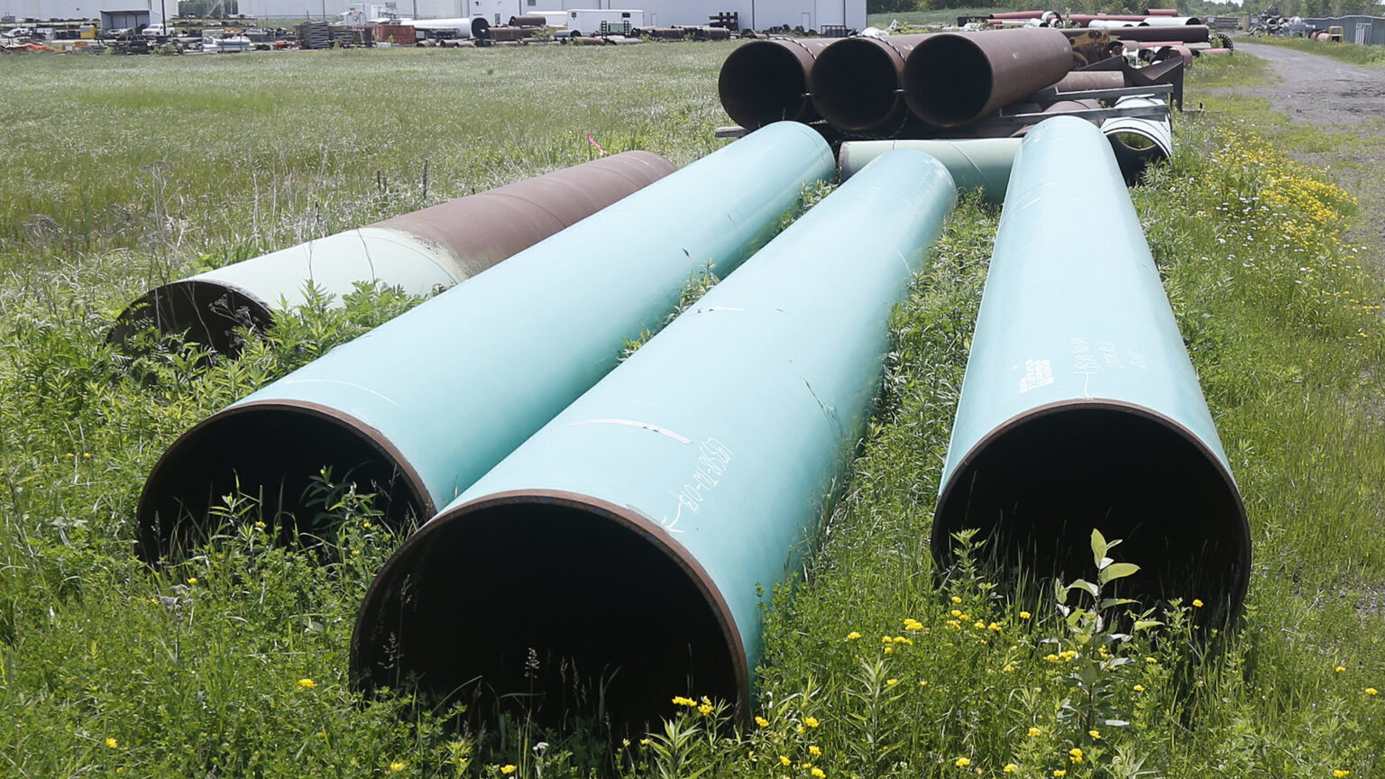 Multiple segments of a metal pipeline sit on an open field, with buildings and industrial equipment in the background.