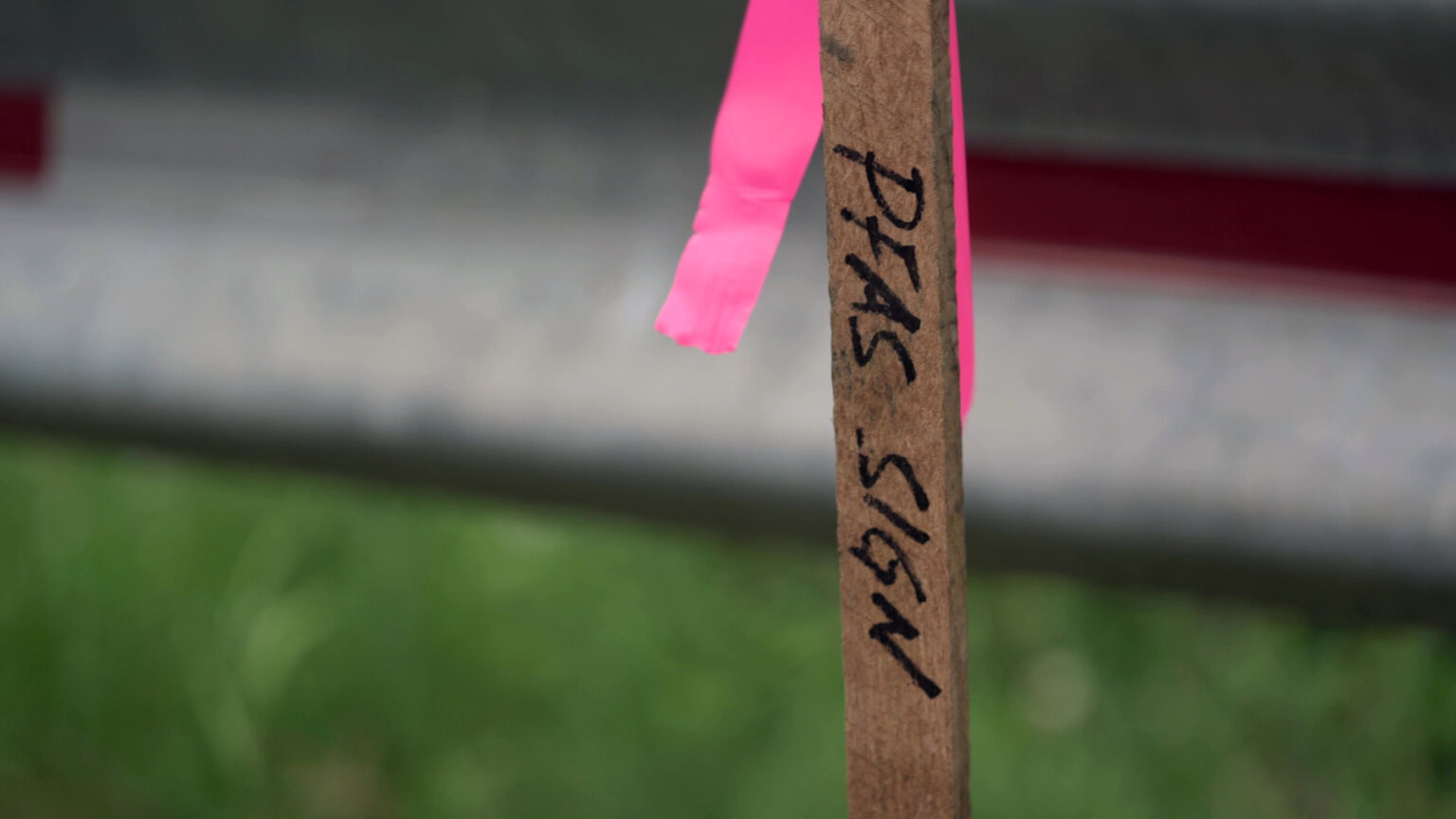 A wooden stake with the words PFAS Sign written in marker stands in front of a metal vehicle barrier.