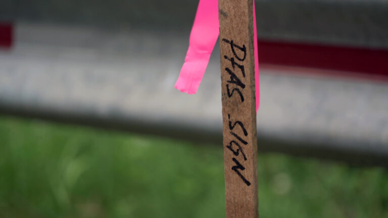 A wooden stake with the words PFAS Sign written in marker stands in front of a metal vehicle barrier.