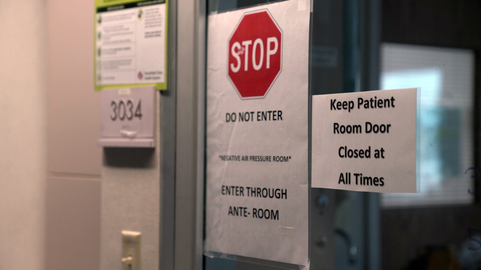 Paper signs reading Stop, Do Not Enter, *Negative Air Pressure Room* and Keep Patient Room Door Closed at All Times at taped to a sliding glass door of a hospital room.