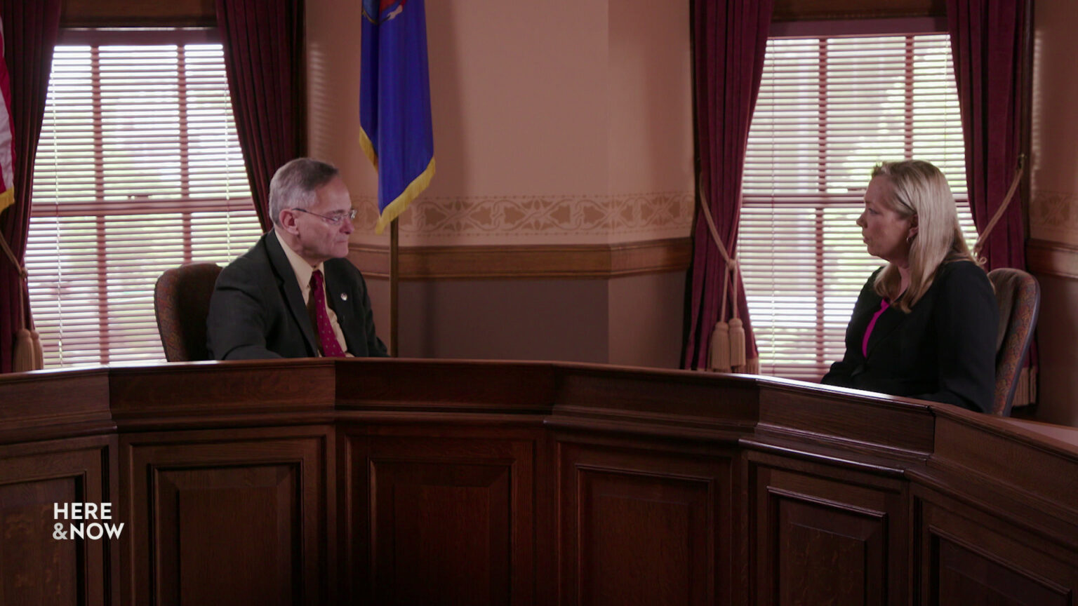 Peter Barca and Frederica Freyberg sit in chairs behind a multi-seat bench-style desk in a room with a Wisconsin flag and windows with lowered open blinds and tied open curtains.