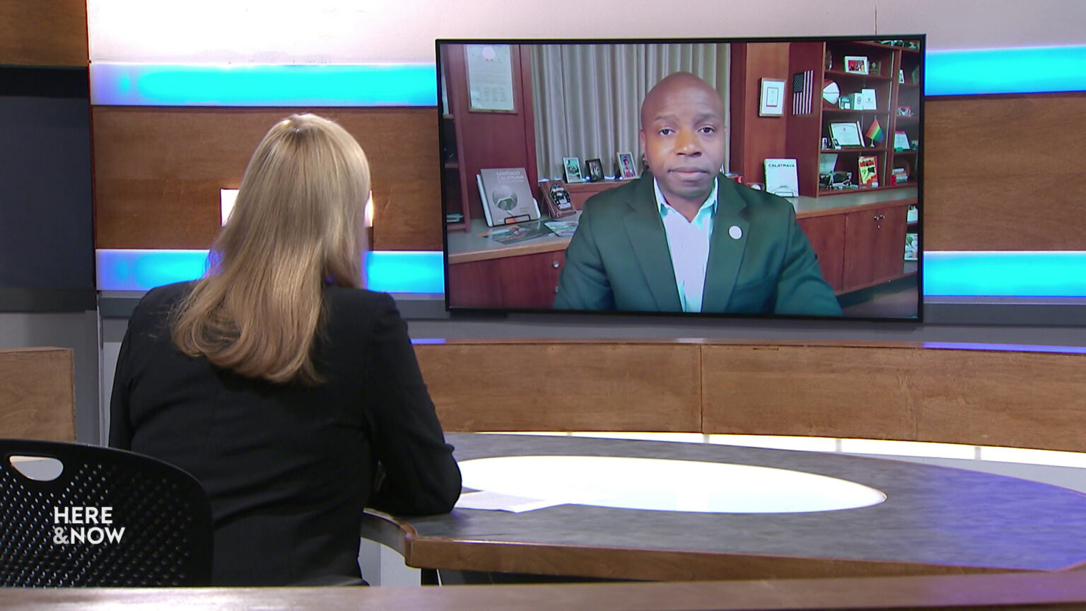 Frederica Freyberg sits at a desk on the Here & Now set and faces a video monitor showing an image of Cavalier Johnson.