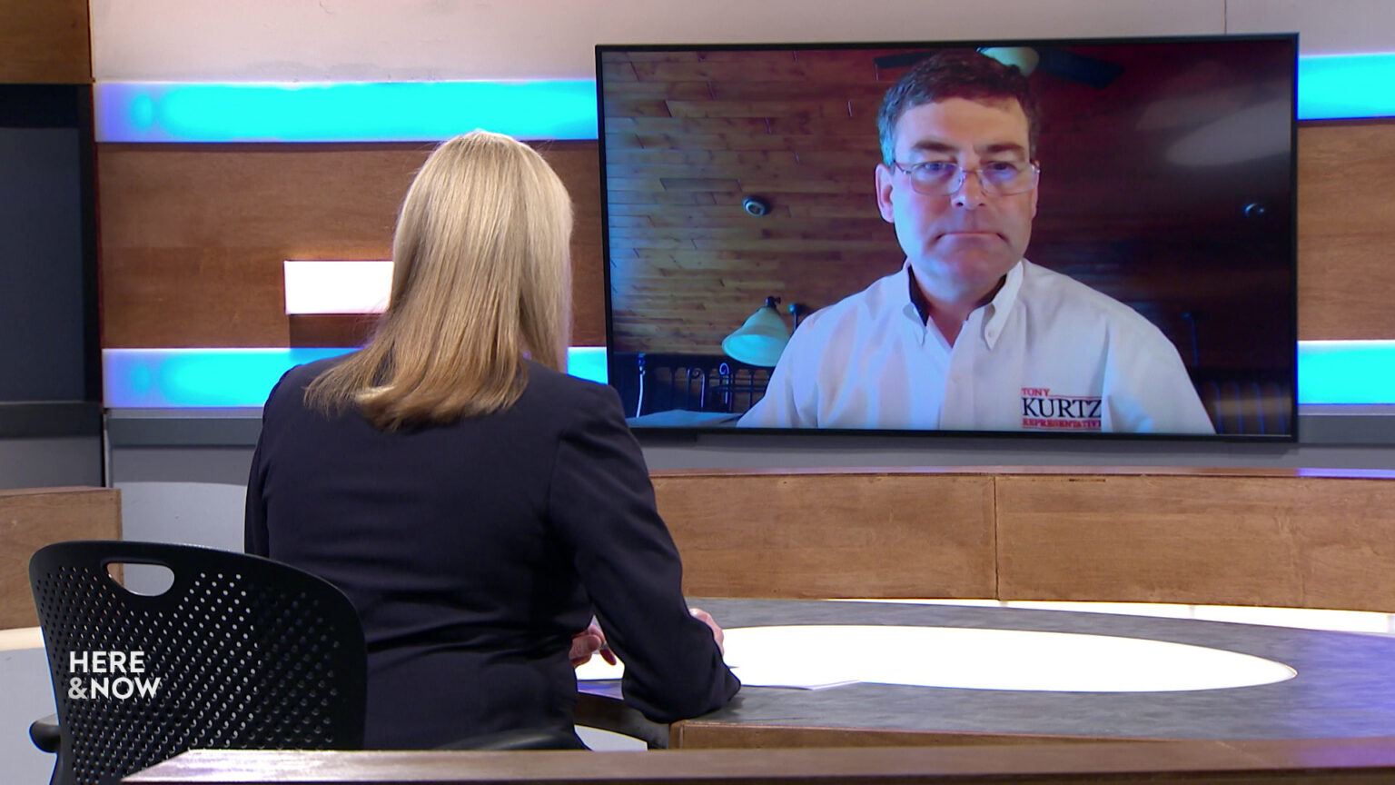 Frederica Freyberg sits at a desk on the Here & Now set and faces a video monitor showing an image of Tony Kurtz.