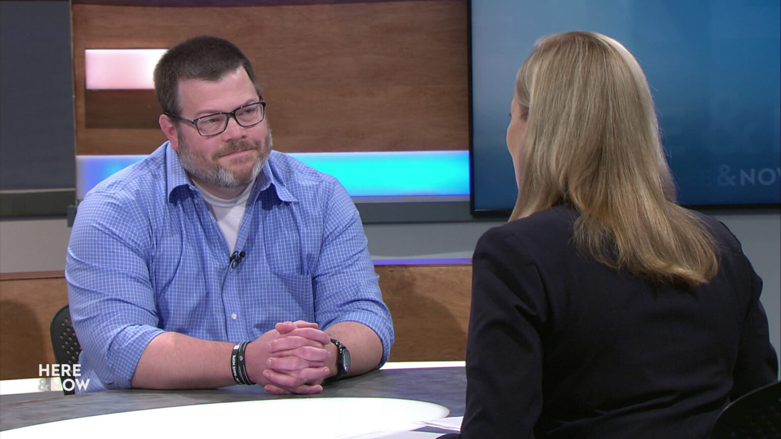 Tony Thrasher and Frederica Freyberg sit facing each other on the Here & Now set.