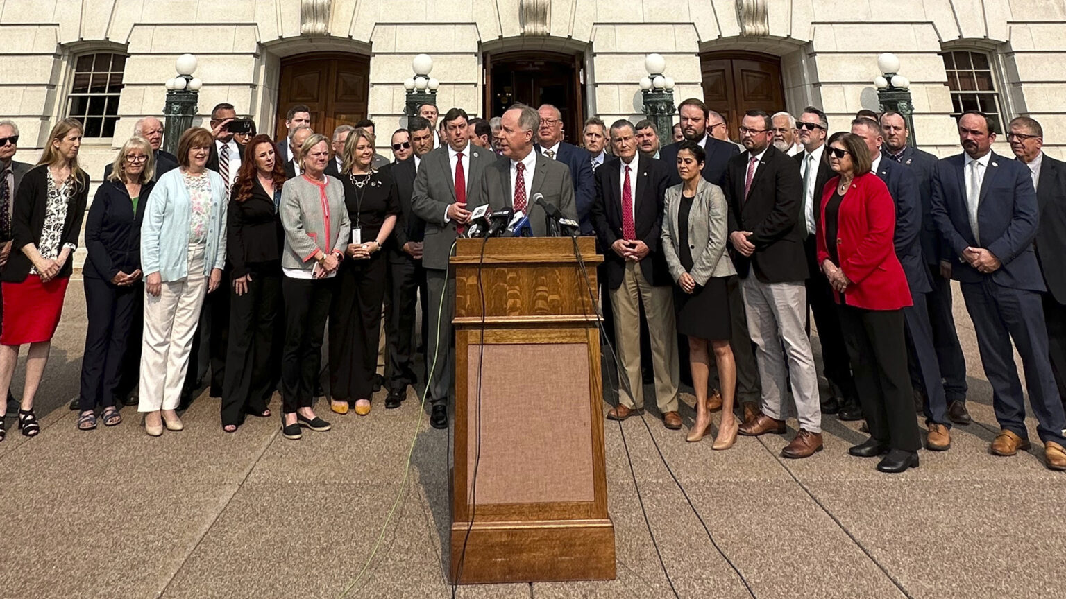 Robin Vos stands behind a wooden podium affixed with multiple microphones with trailing cords, with dozens of people standing behind him on a plaza outside an entrance to the a building with marble masonry and art deco bronze light fixtures.
