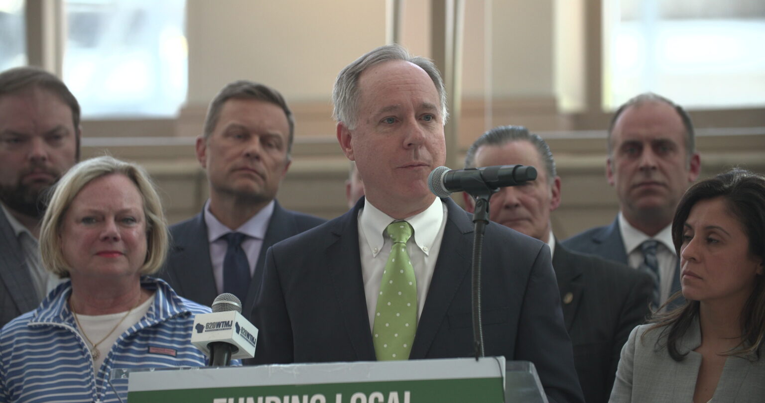Robin Vos stands at a podium and speaks into a microphone, with other people standing beside and behind him.
