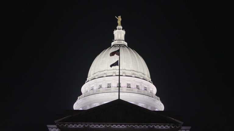 The U.S. and Wisconsin flags wave on a flagpole atop the roof of one wing of the Wisconsin Capitol Building at night, silhouetted against the illuminated dome topped by a statue.
