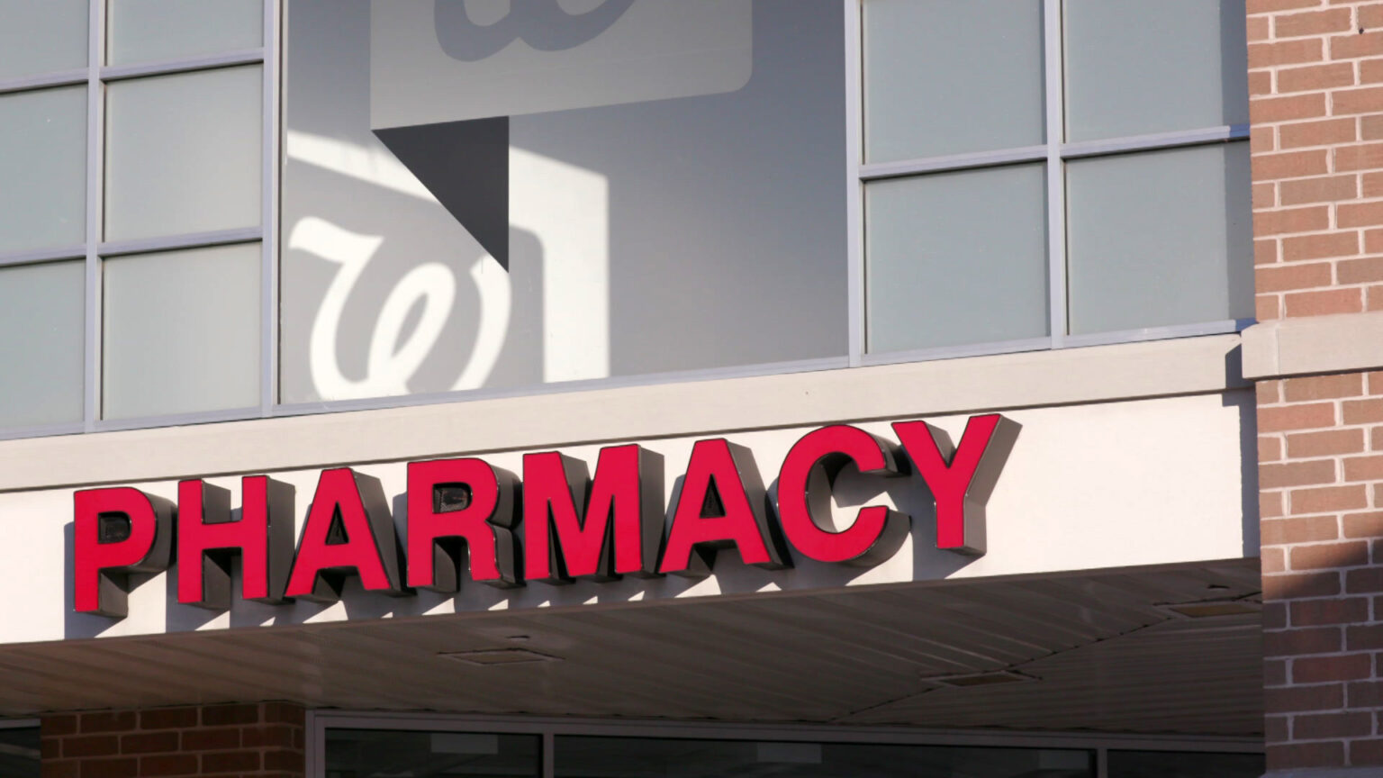 Sign letters affixed to an awning above the entrance to a building spell PHARMACY, with the shadow of a window label of the cursive w wordmark for Walgreens visible above.