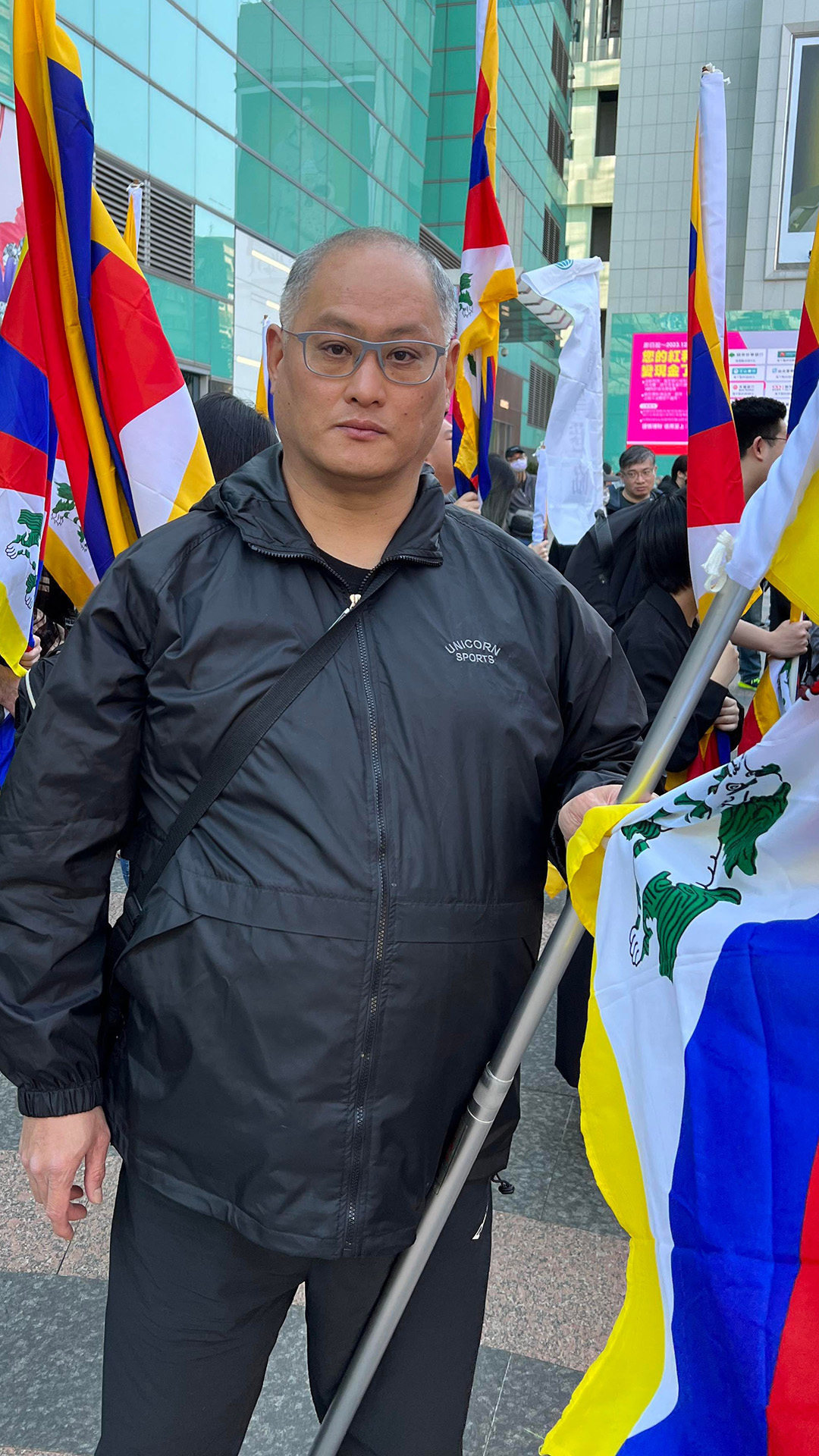 Lee Ming-che stands and holds a flag in an outdoor plaza alongside other people holding flags, with buildings in the background.
