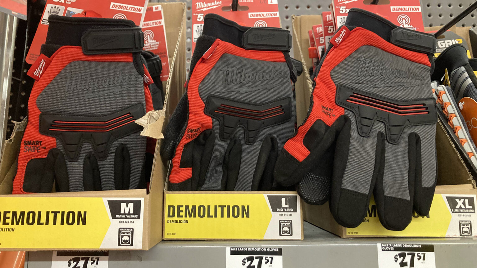 Three different sizes of work gloves with knuckle pads and Velcro wrist-fasteners are arranged on hangers and in boxes on a shelf, with prices on stickers beneath each.