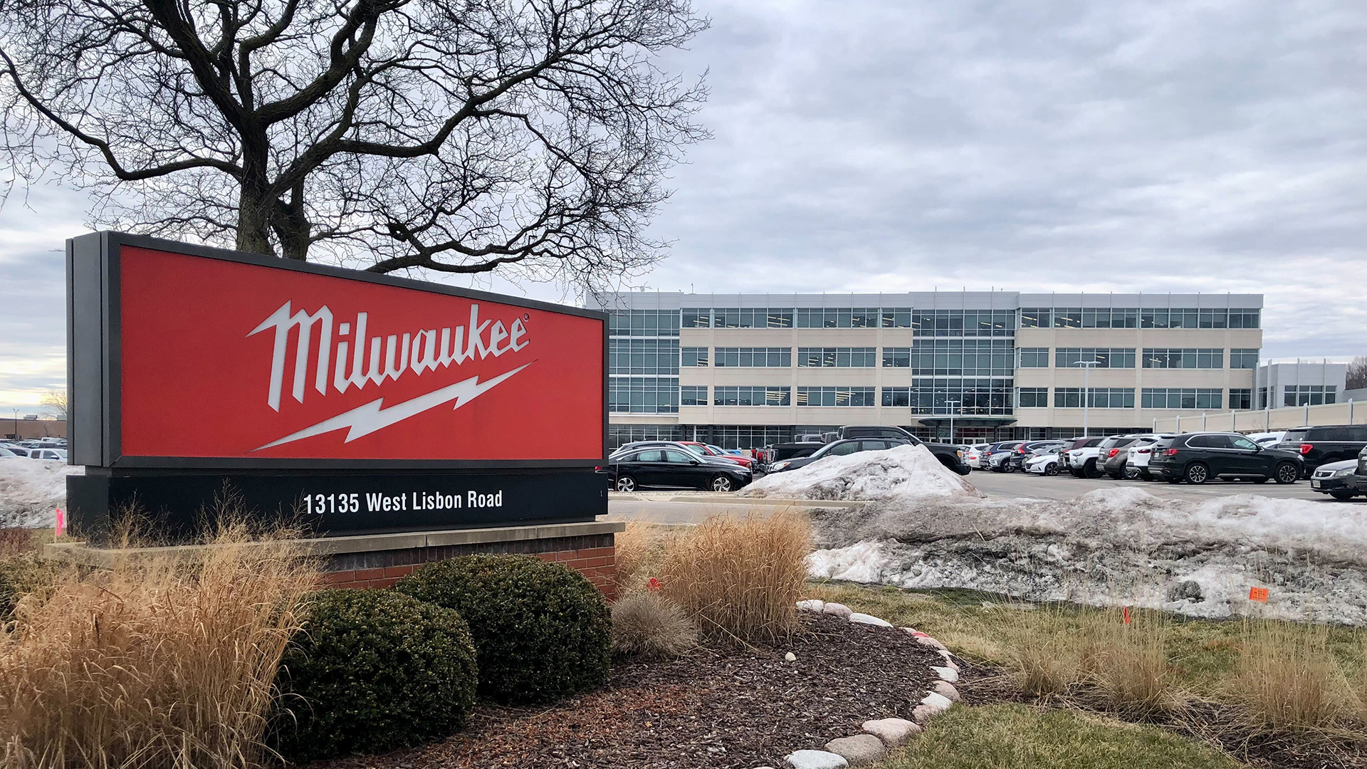 A sign with the Milwaukee Tool wordmark and lightning bolt logo is surrounded by bushes near the entrance to a parking lot, with a tree, parked cars and a multi-story office building in the background.