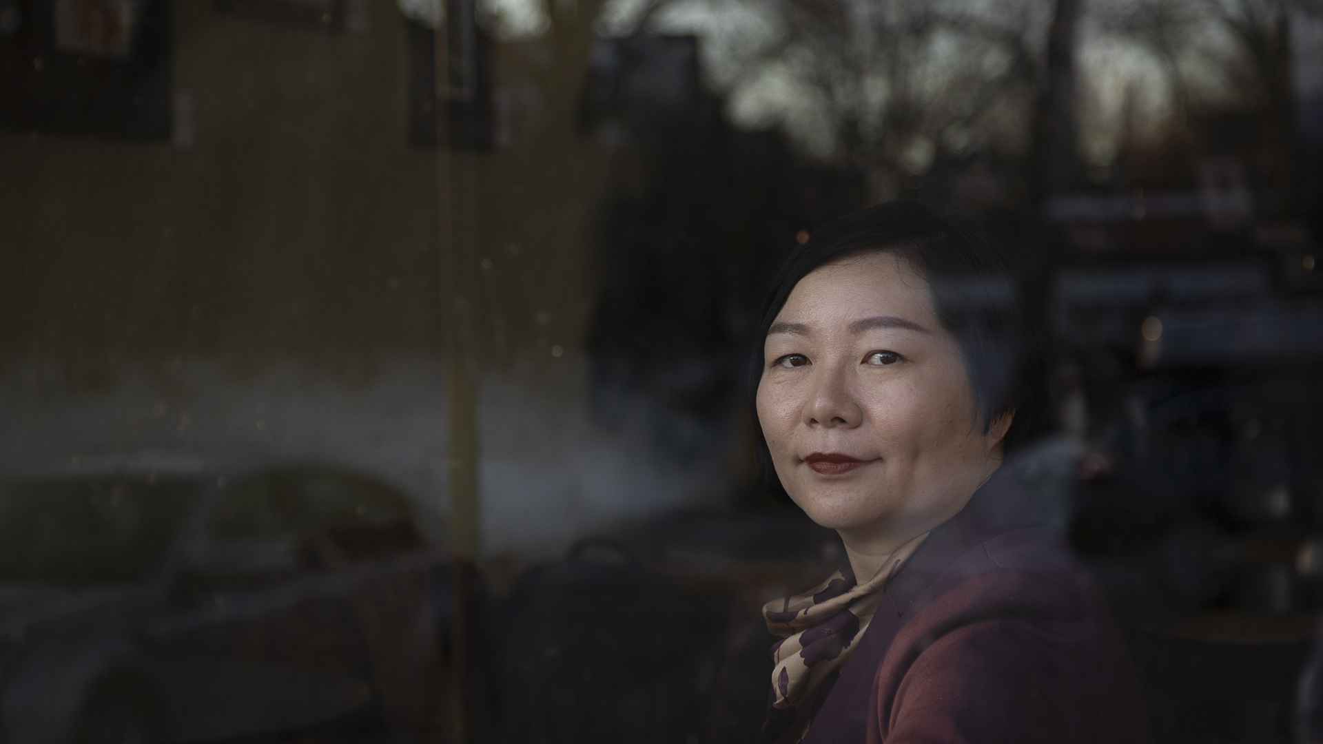 Shi Minglei sits inside a building and looks through a large plate-glass window, with parked cars and trees visible in its surface reflection.