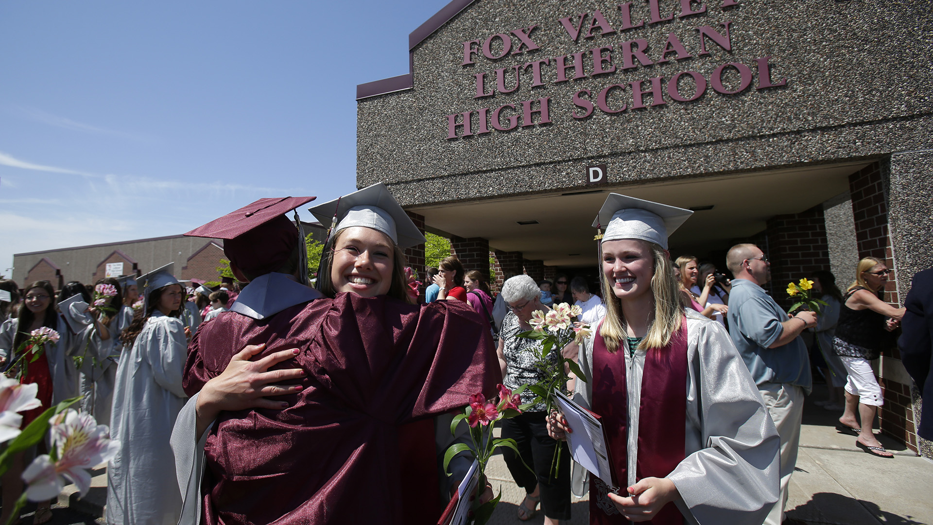 Two students wearing graduation robes and caps hug each other with other graduates and other family members standing on a sidewalk in front of a building with a pebble façade with a sign that reads "Fox Valley Lutheran High School."