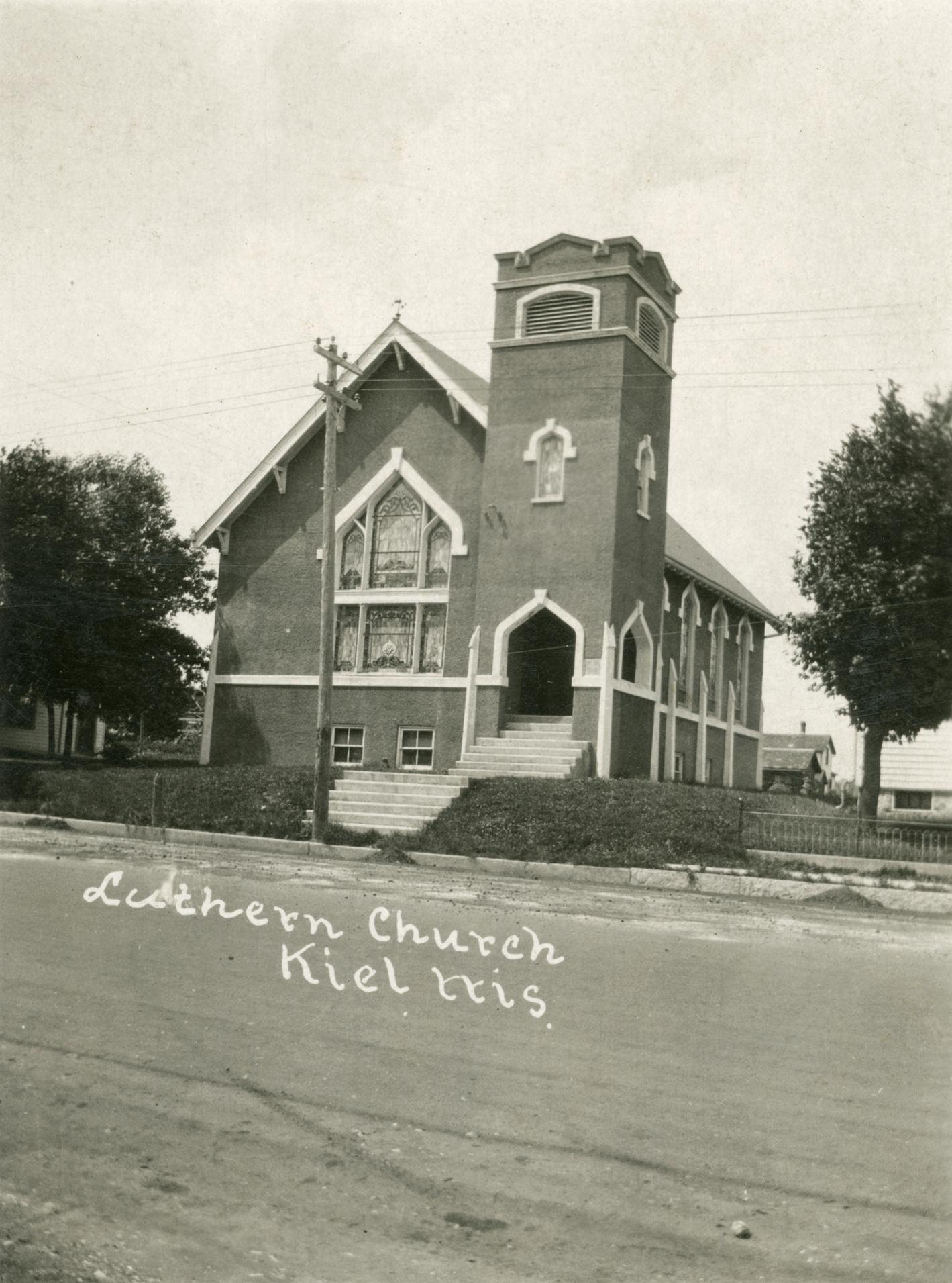 A sepia-toned historic photo shows a brick church building with a bell tower standing along the side of a road between two trees, with handwritten script on the image reading "Luthern Church" and "Kiel Wis."