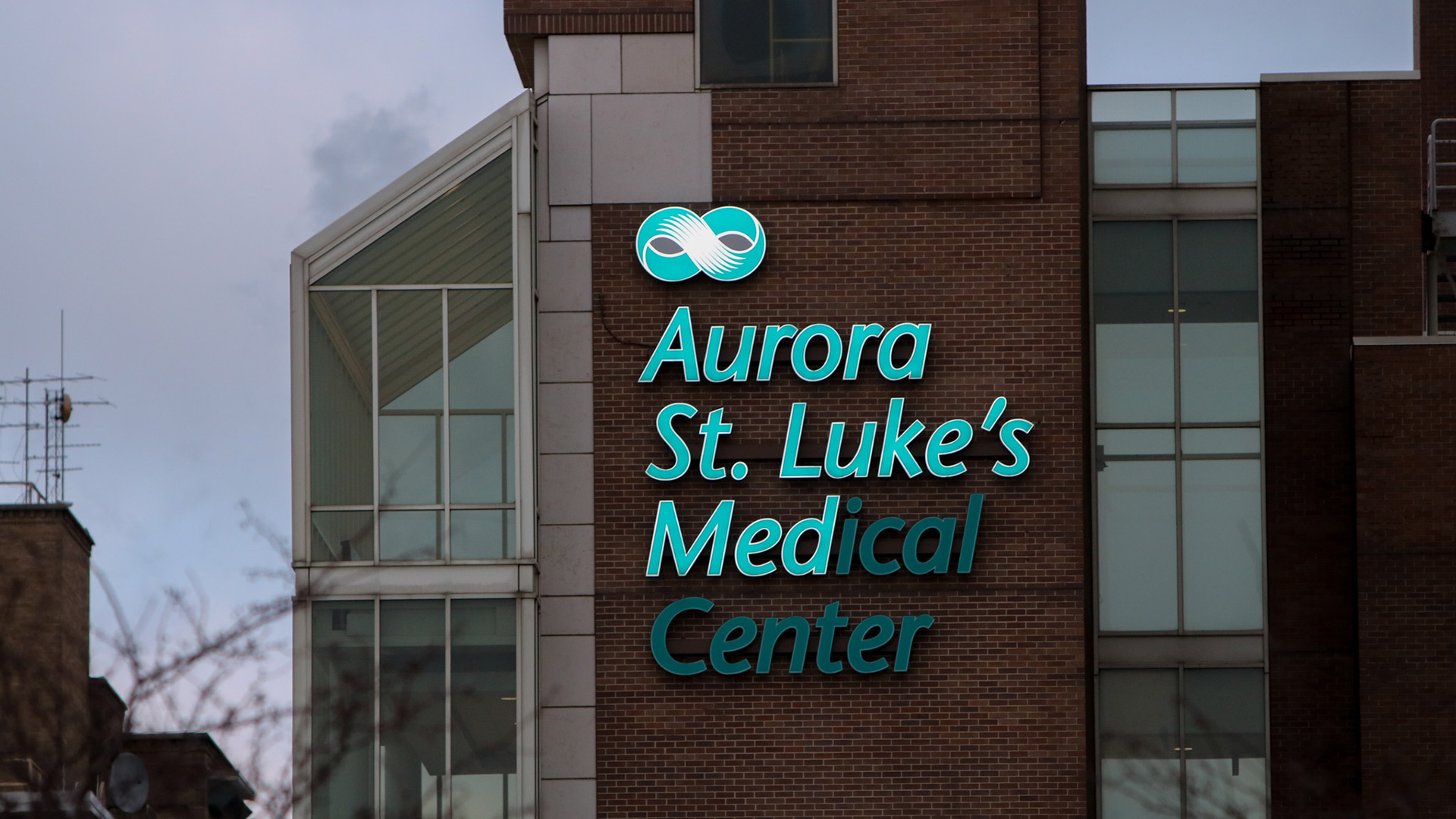 A multi-story building with glass-walled walkways and brick elevator shafts includes a letter sign near its roof that reads "Aurora St. Luke's Medical Center."