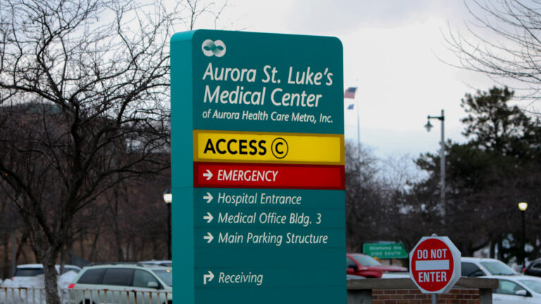 A parking lot sign for Aurora St. Luke's Medical Center of Aurora Health Care Metro, Inc. includes arrows and signs pointing toward access for Emergency, Hospital Entrance, Medical Office Bldg. 3, Main Parking Structure, and Receiving needs, with parked cars and trees in the background.