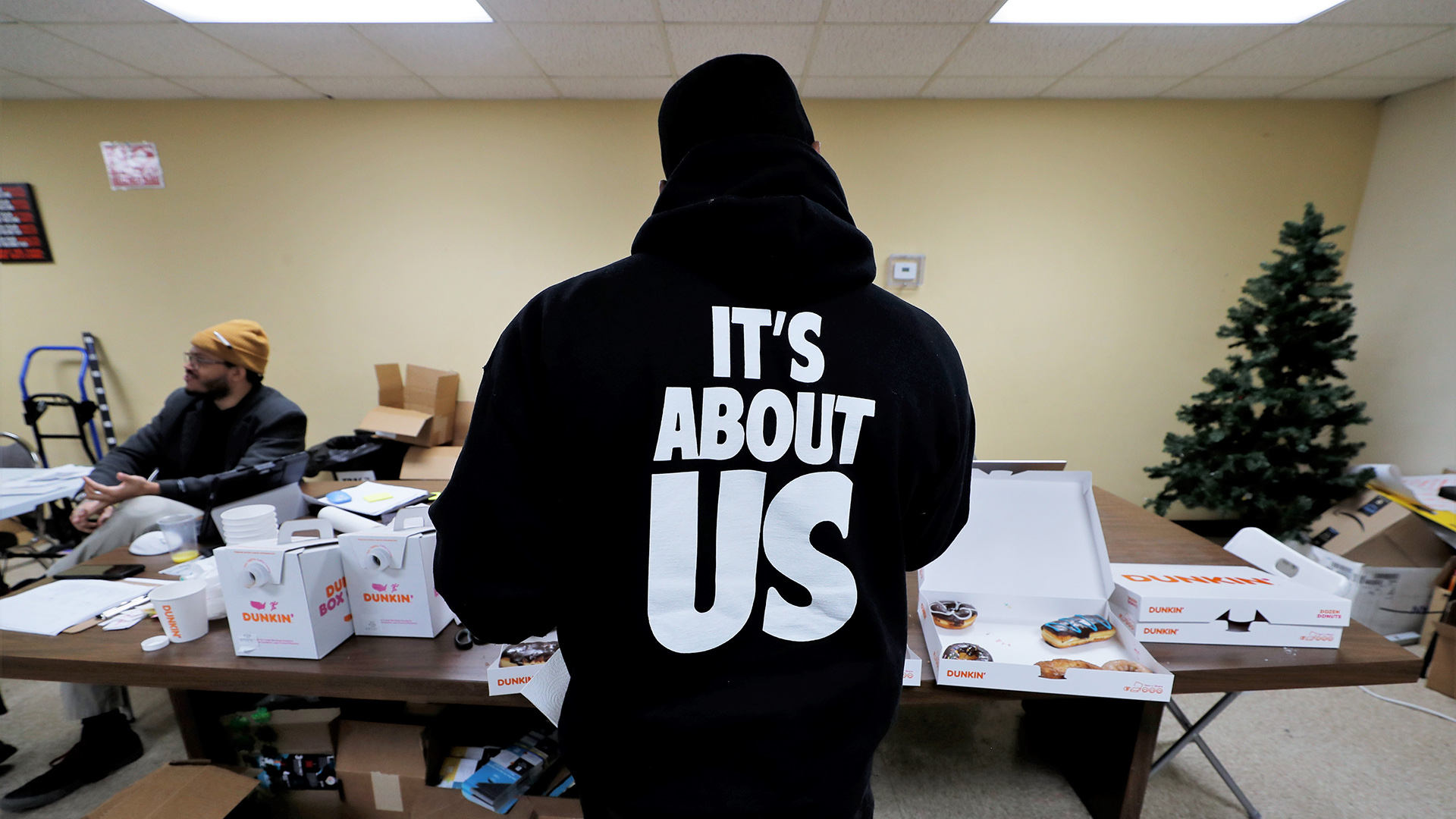 A person wearing a shirt with the words "It's About US" stenciled on the back stands and faces folding tables stacked with boxes of donuts and coffee, with another person seated to the side in a room with stacked boxes and an artificial Christmas tree.